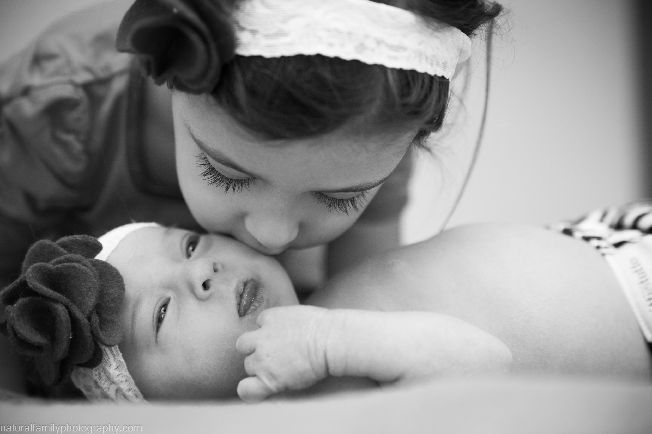 A woman is kissing a newborn baby girl on the forehead while lying in a hospital bed. The baby is wearing a headband with dark decorative flowers, and she is looking at the camera with her hand near her face.
