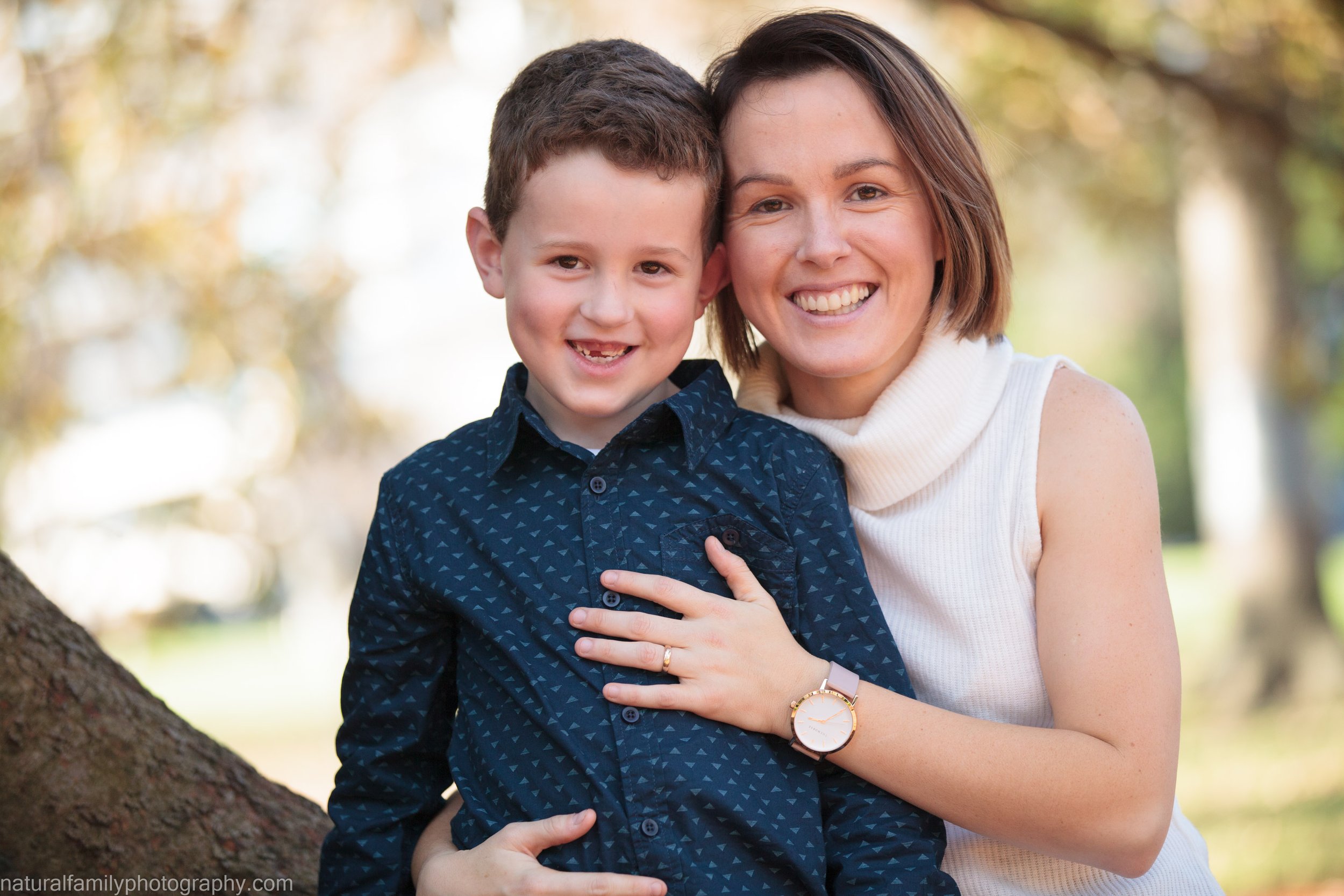 A smiling woman and young boy outdoors in a park with autumn trees in the background, hugging each other.
