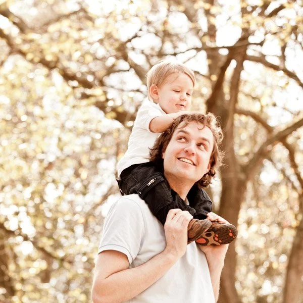 A young man carrying a toddler on his shoulders outdoors in a park with trees and sunlight. Candid portrait of a father and son by Natural Family Photography, Melbourne.