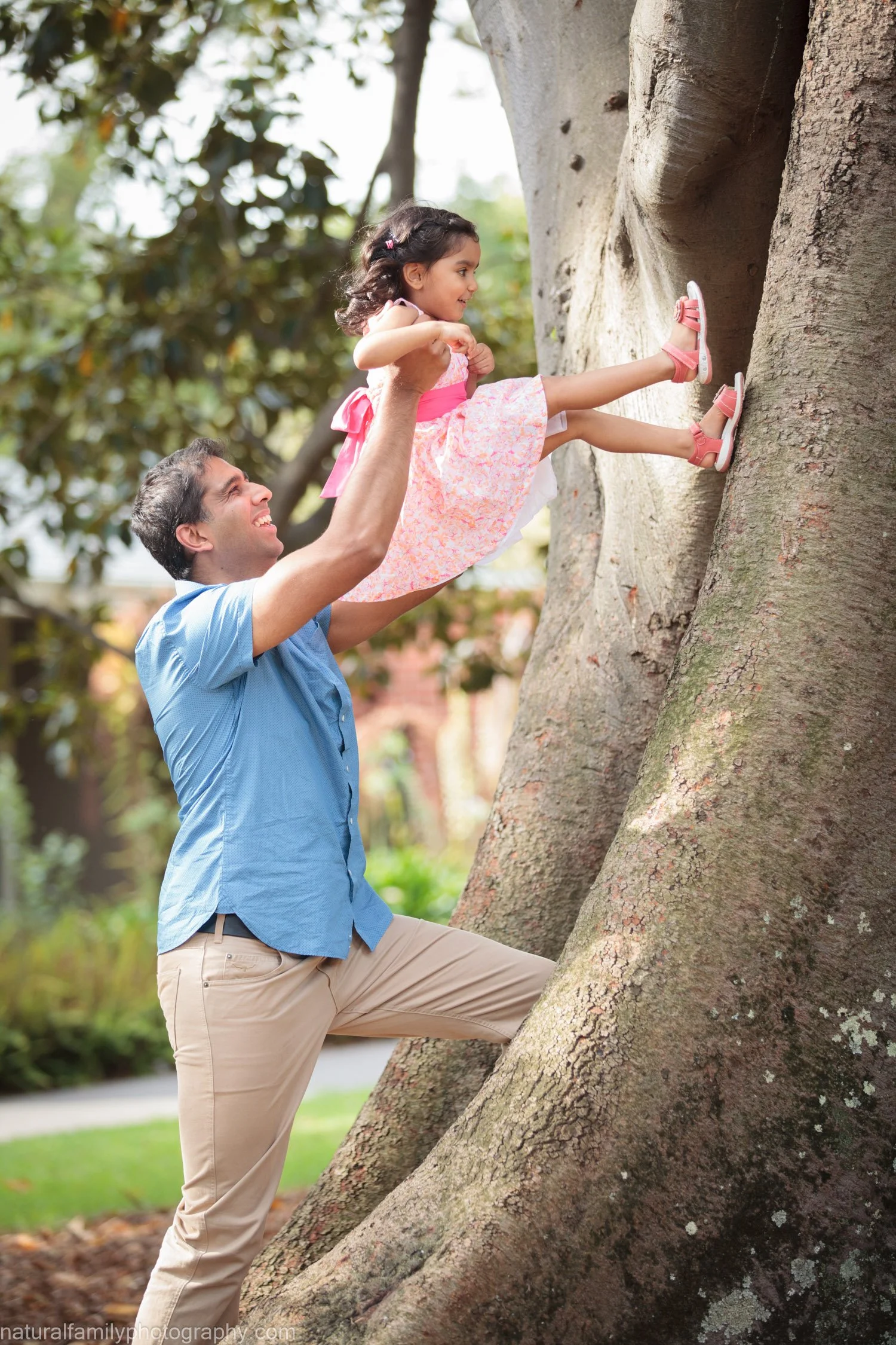 A man helping a young girl climb a large tree outdoors in a park, with greenery in the background.