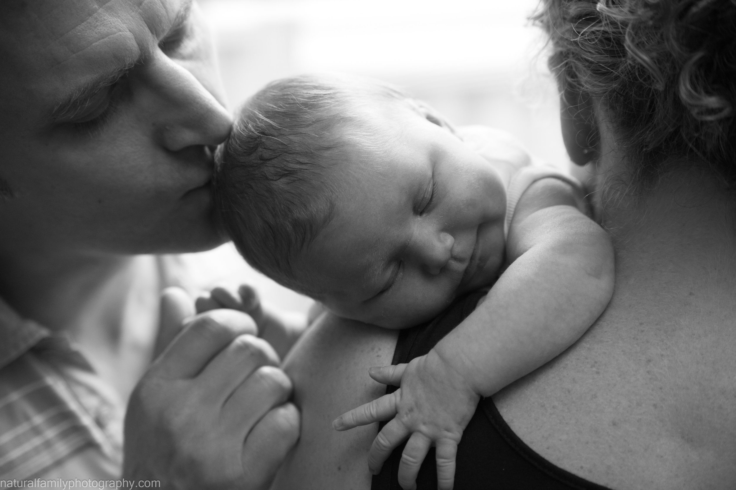 Black and white photo of a newborn baby resting on a woman's shoulder, with a man kissing the baby's head, all close together.