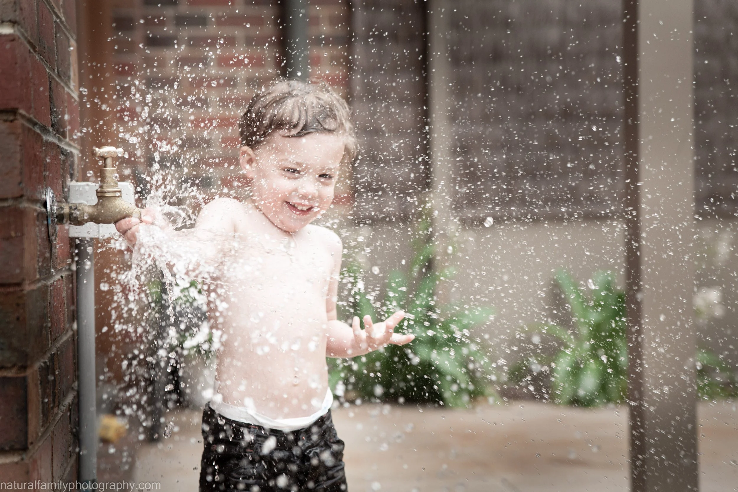 Young boy playing in water spraying from a garden hose on a brick wall, smiling and with his arms outstretched, enjoying outdoor fun.