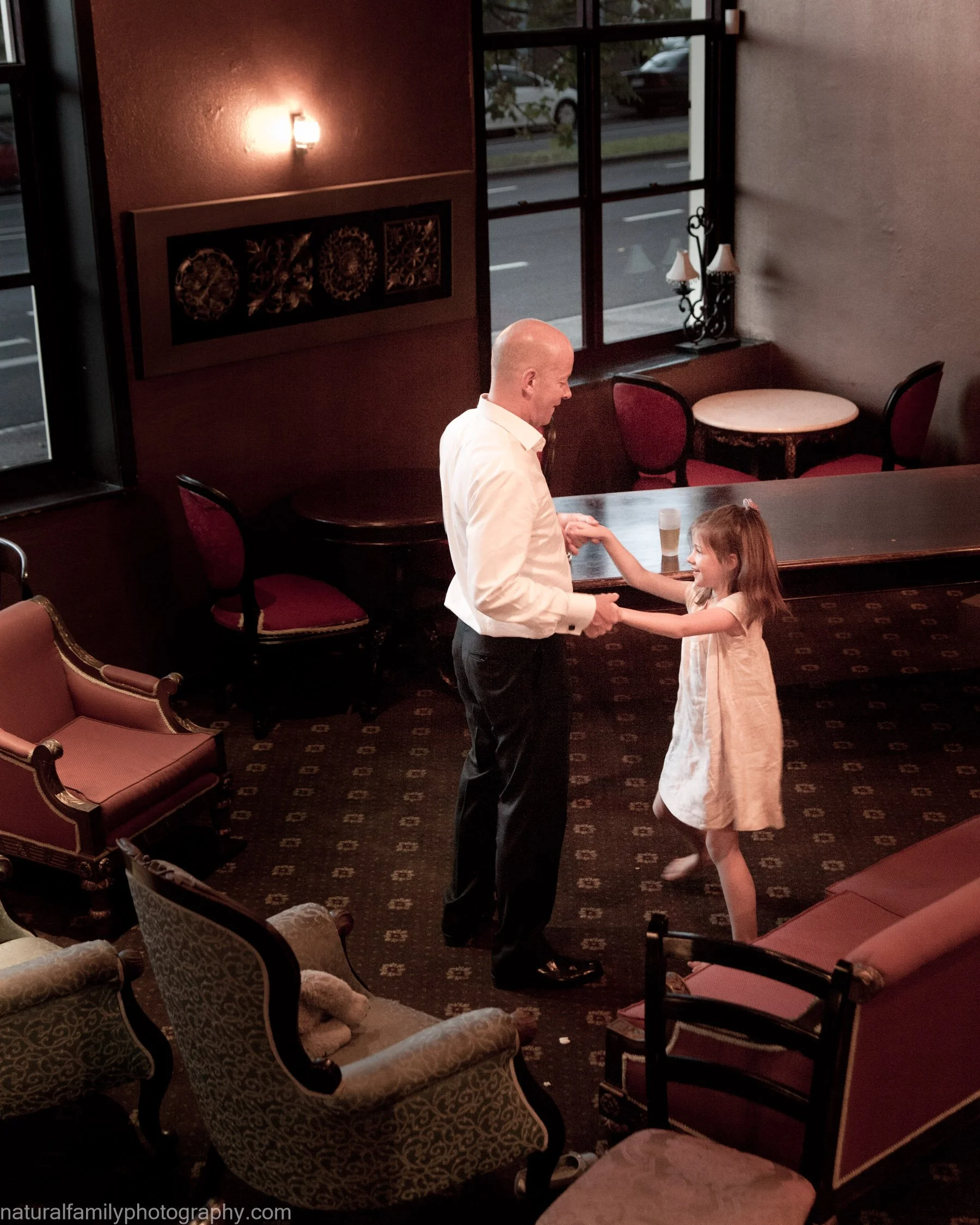 A man and a young girl are dancing and holding hands in a warmly lit restaurant or lounge. The man has a bald head and is wearing a white dress shirt and black pants. The girl is in a white dress and has brown hair. There are upholstered chairs, tabl