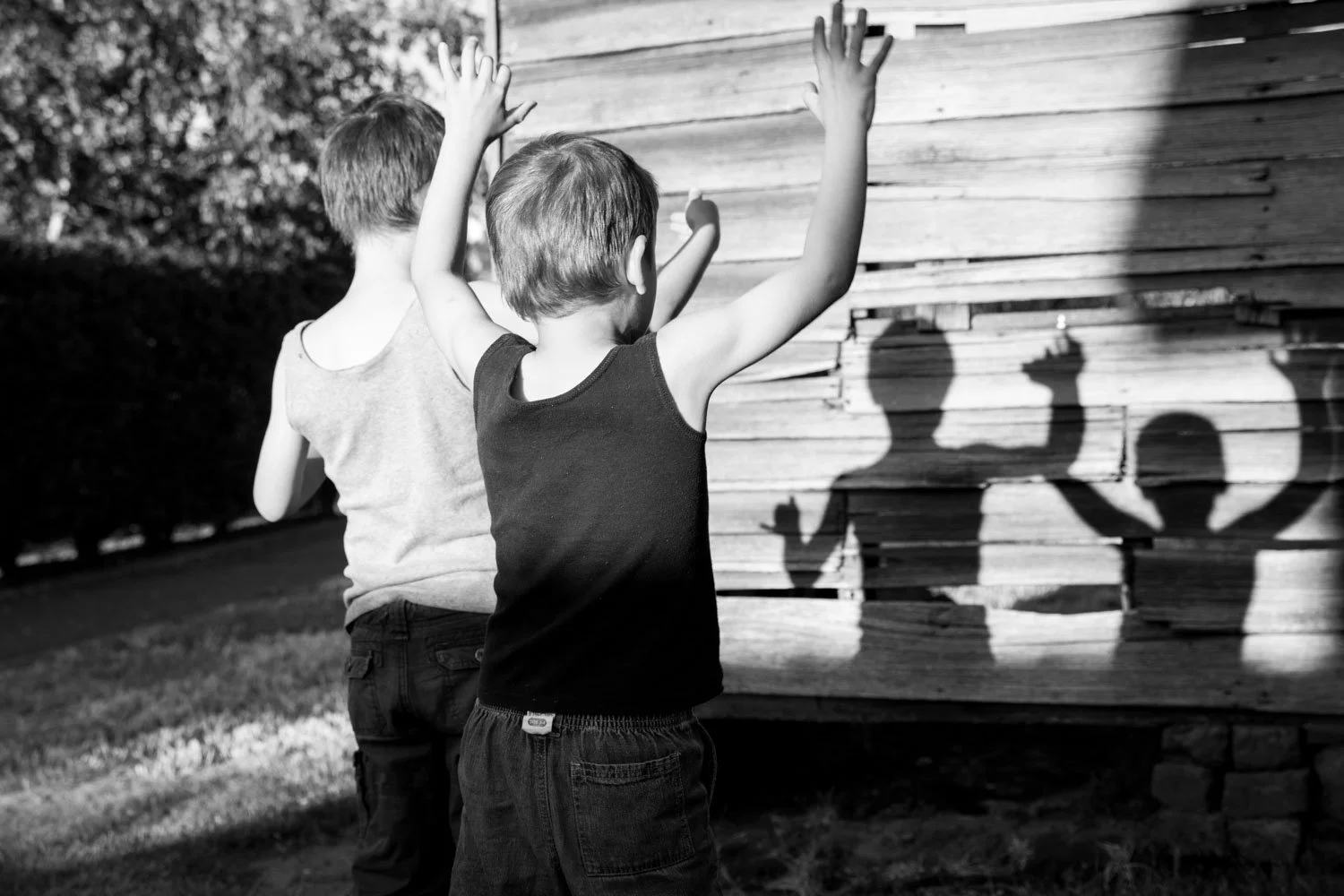 Two young boys are playing outside, casting shadows on a wooden wall, with one boy raising his arms and the other slightly turned away.