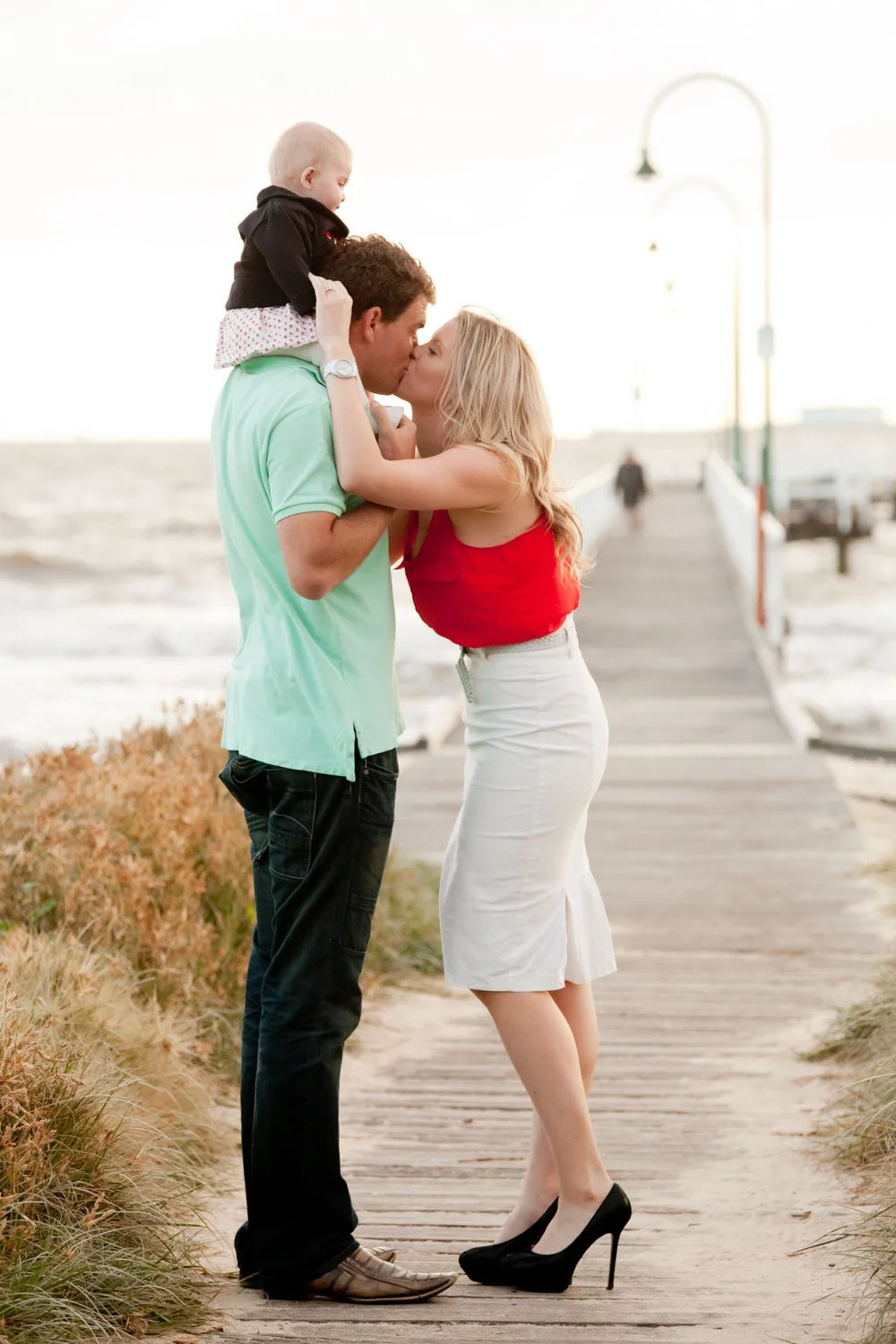 A couple sharing a kiss on a wooden pier on a beach with a small child on the man's shoulders, holding his face, during sunset.