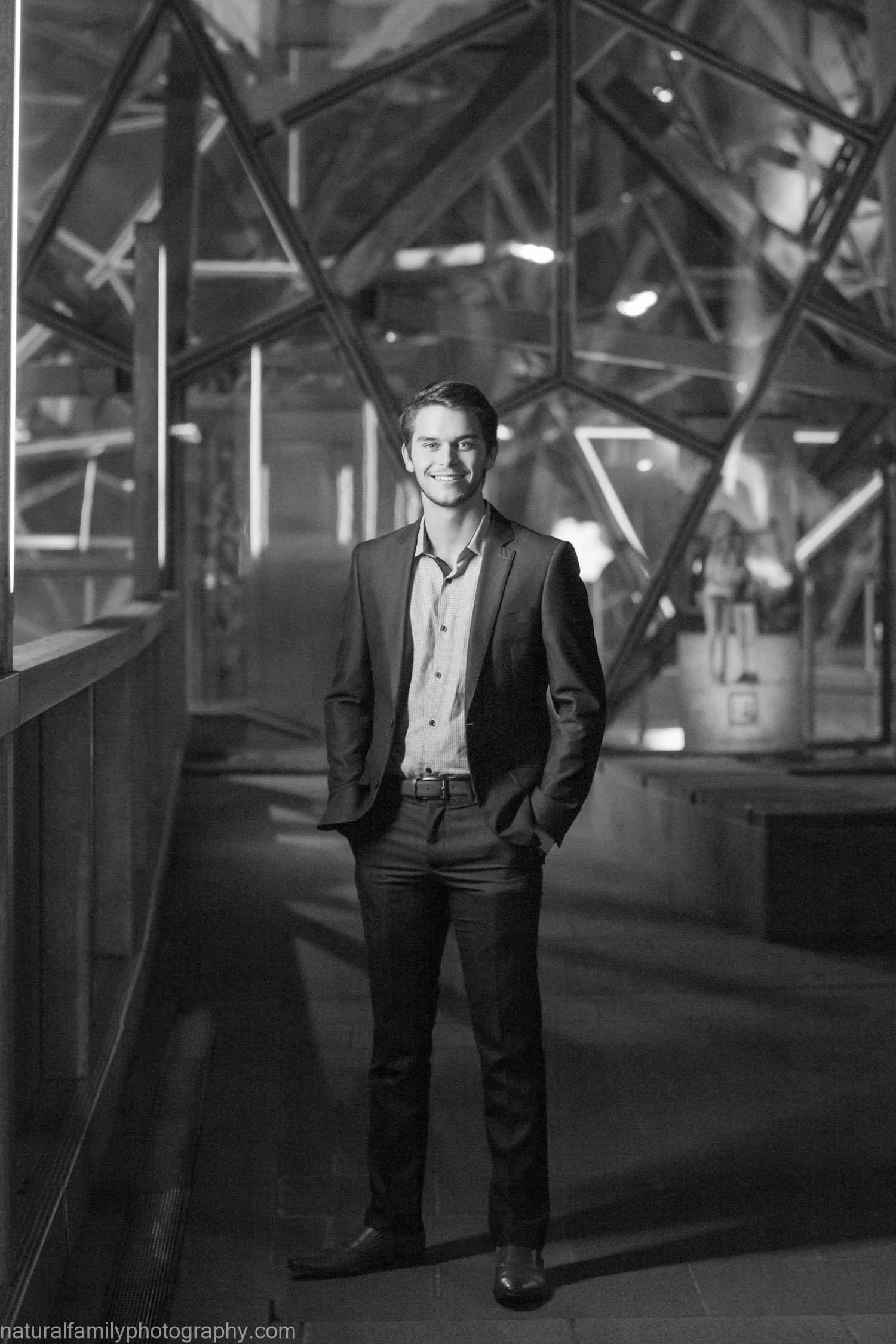 A young man in a suit standing with his hands in his pockets inside a modern building with geometric glass architecture in the background.