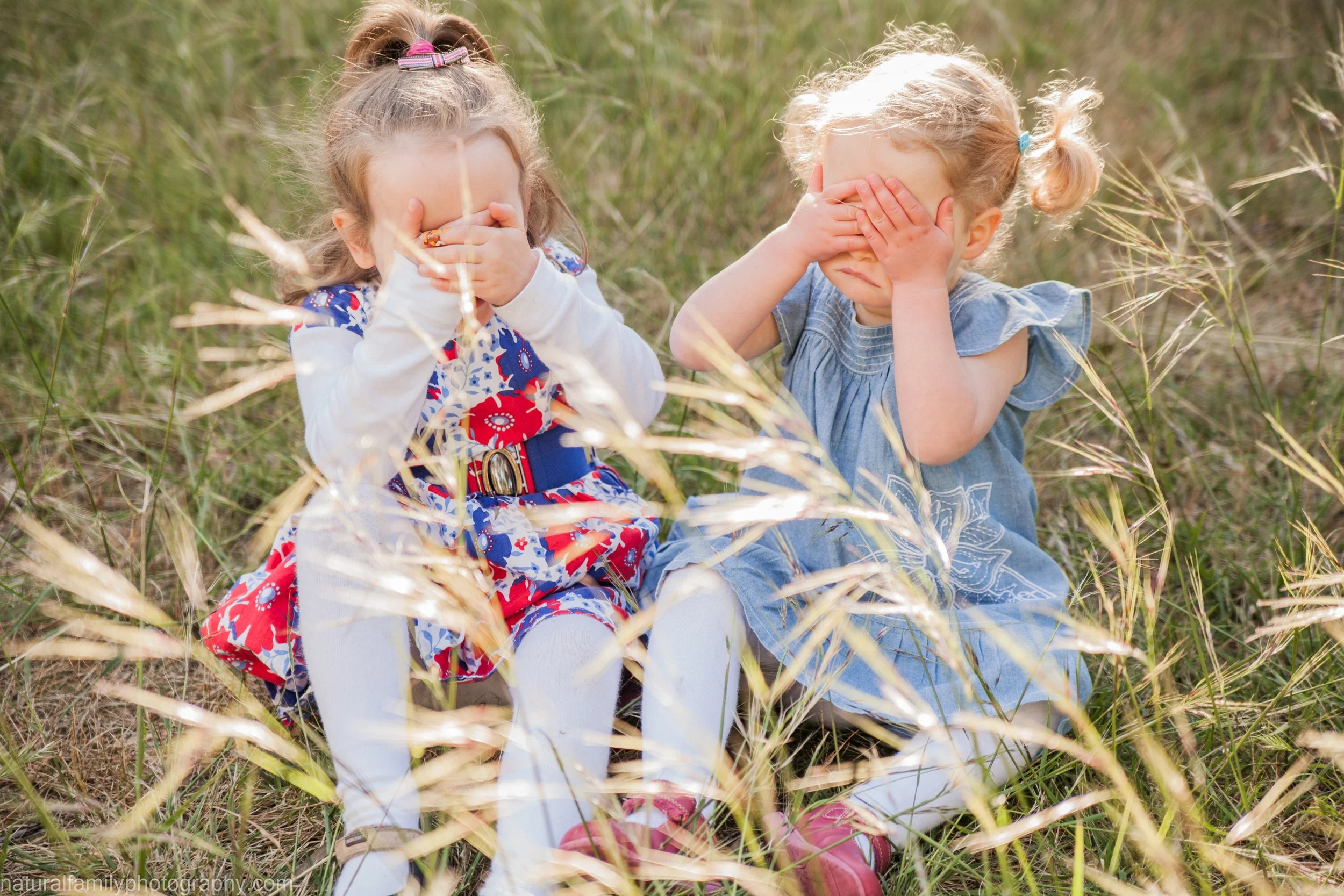 Two young girls sitting in a grassy field covering their eyes with their hands. Classic children's portrait by Natural Family Photography, Melbourne.