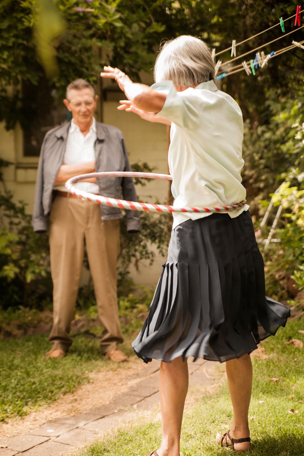 An elderly woman hula hooping in her backyard while an elderly man watches her, standing with his arms crossed and smiling.
