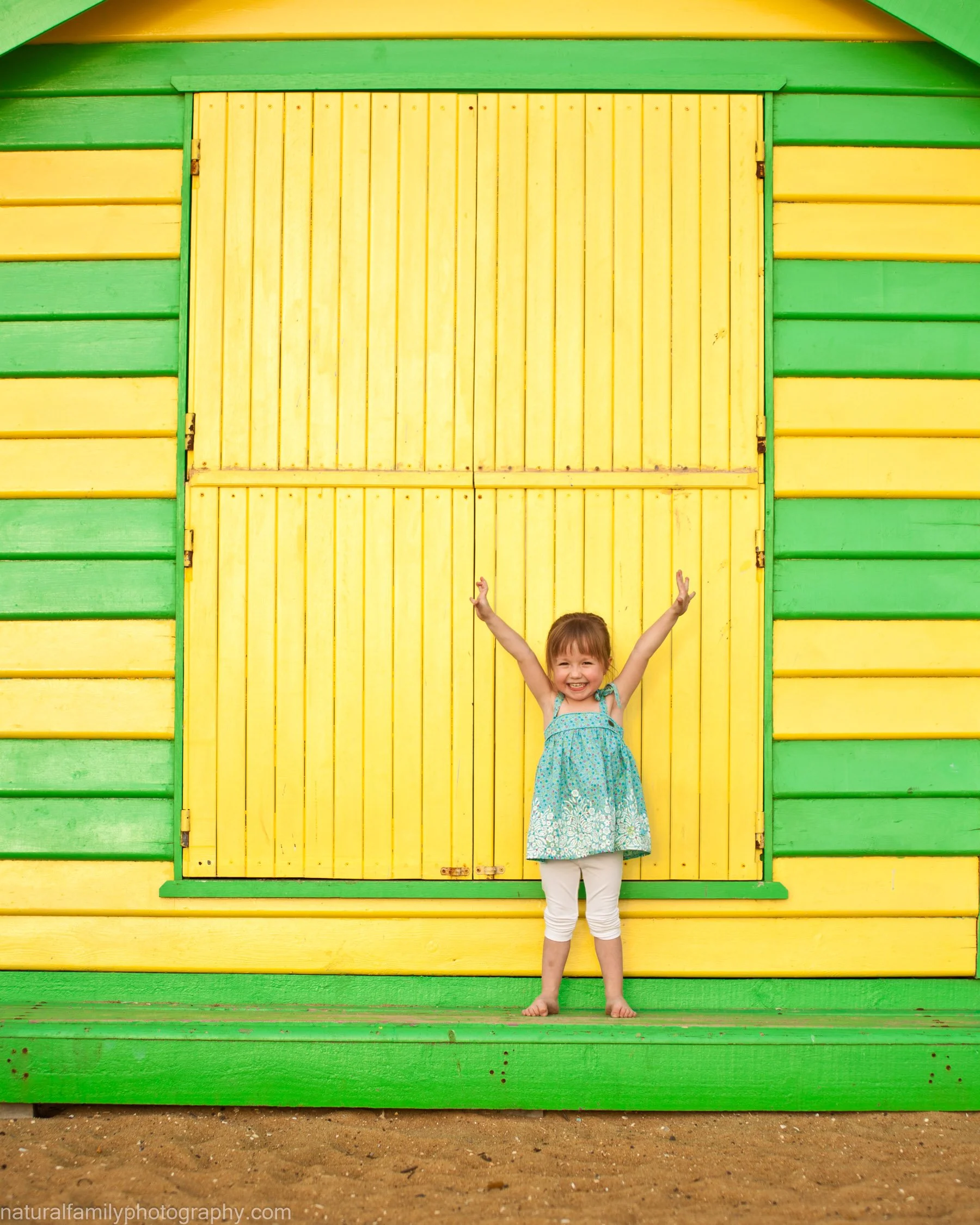 A young girl with brown hair in a blue dress and white leggings standing in front of a colourful wooden beach box in Brighton with yellow doors and green trim, smiling and raising her arms in joy. Portrait by Natural Family Photography, Melbourne.