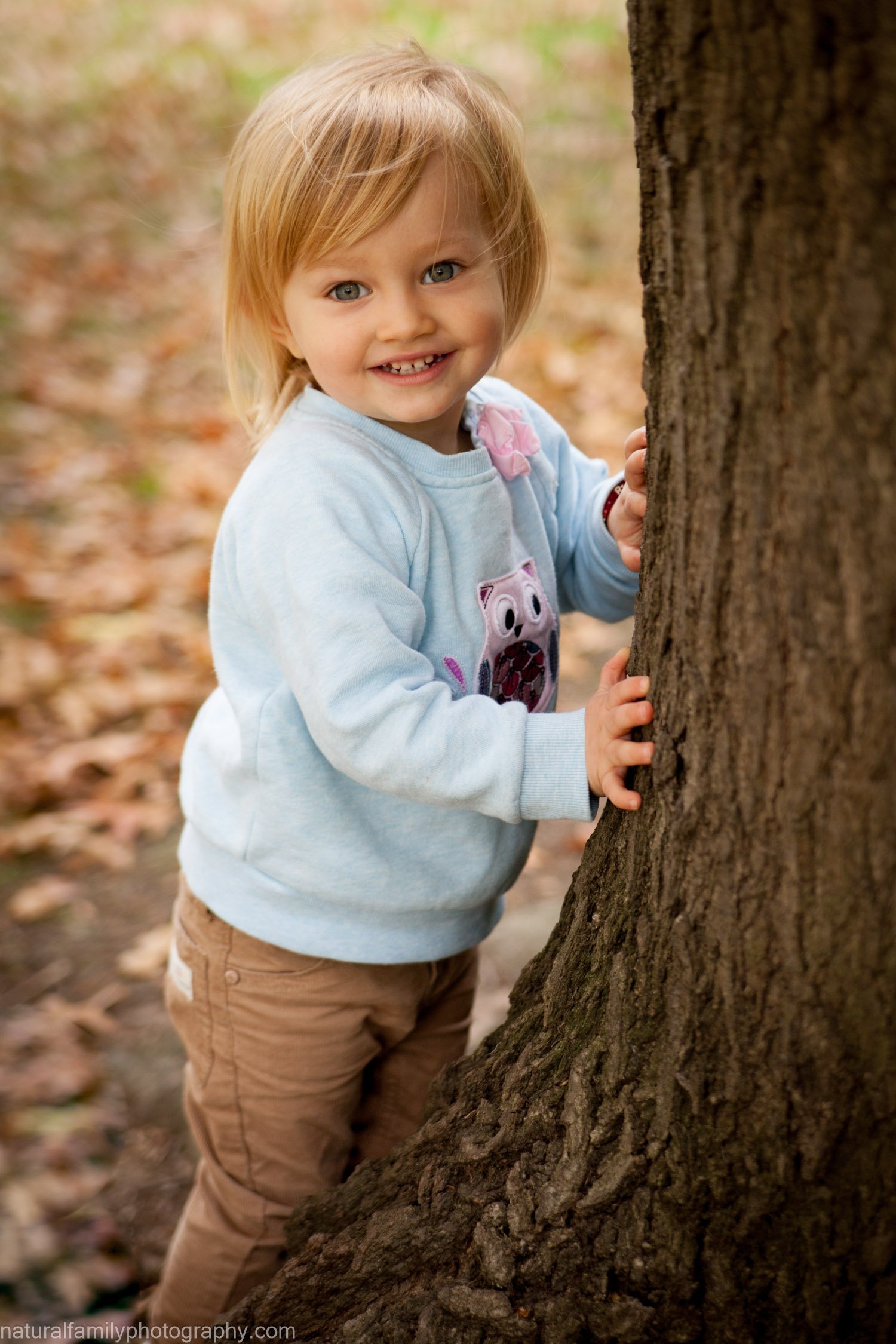 A young girl with blonde hair, blue eyes, and a bright smile, wearing a light blue sweatshirt with a pig design, and tan pants, standing outdoors in a wooded area, holding onto a tree trunk with both hands.