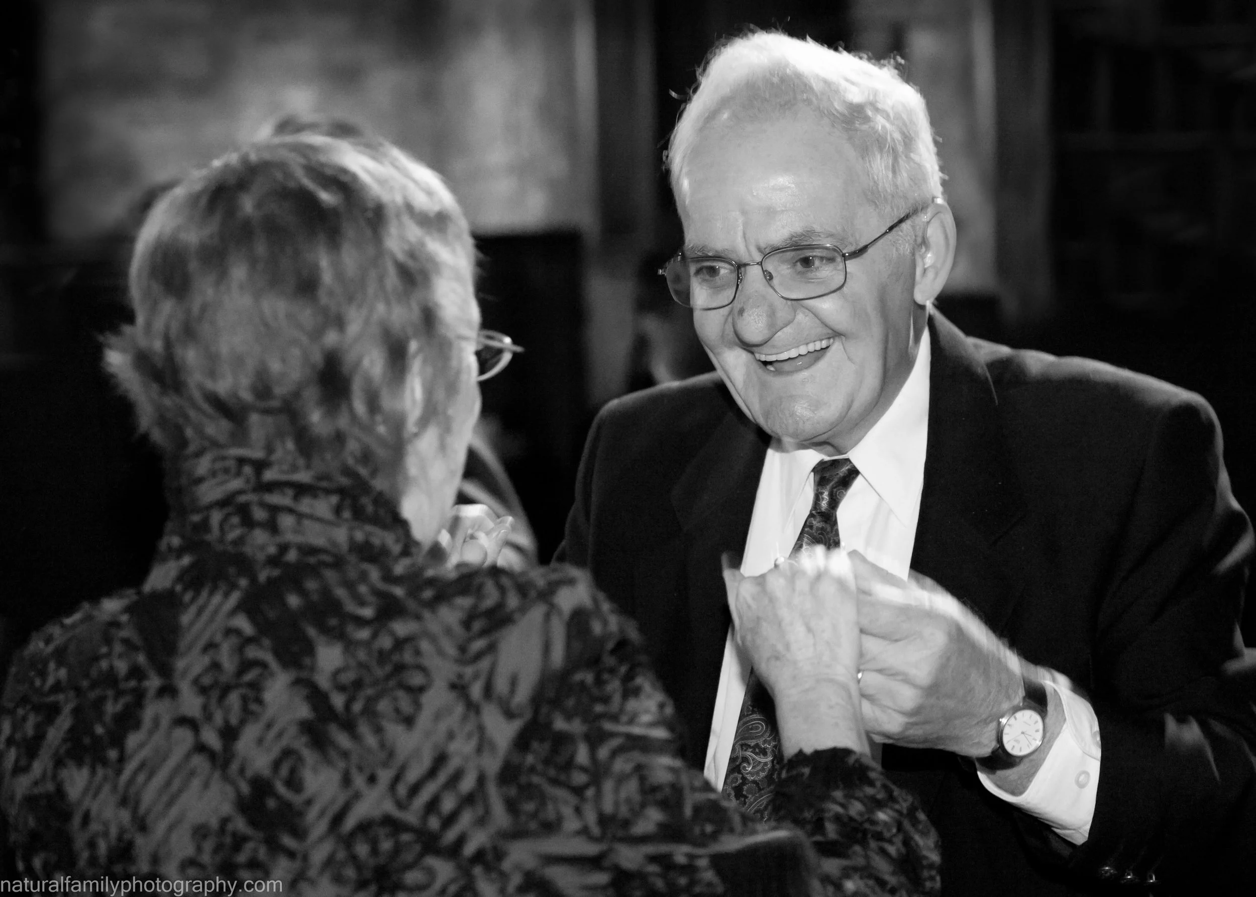 An elderly man in a suit and glasses smiling while holding hands with an elderly woman wearing glasses and a patterned top, indoors. CLassic black and white event image by Natural Family Photography, Melbourne.