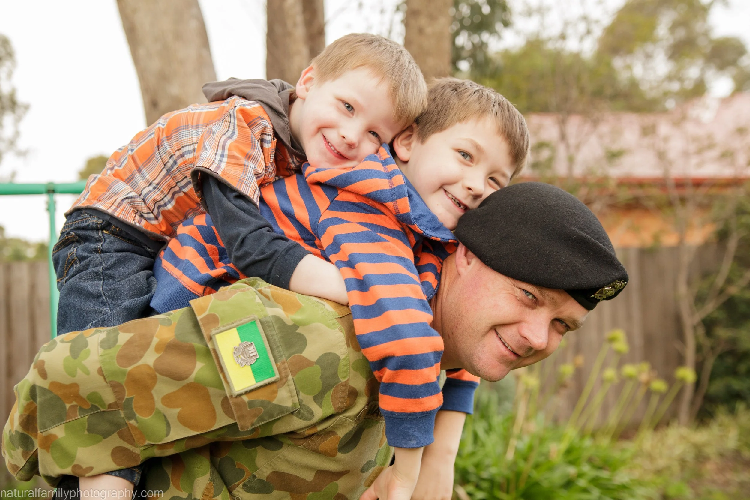 A soldier with a black beret carrying two young boys on his back, outdoors with trees and a fence in the background.