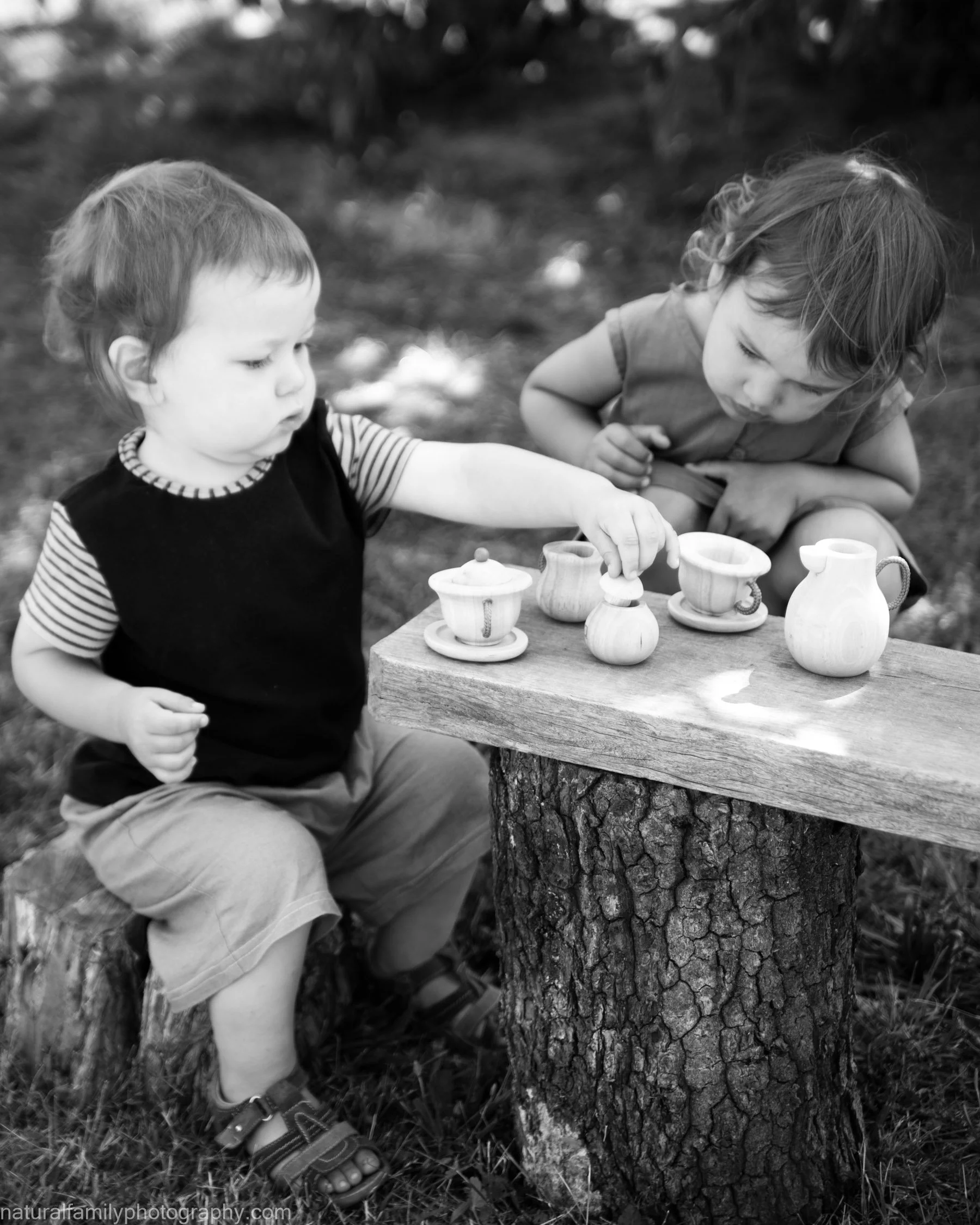 Two young children playing with toy kitchenware on a wooden table outdoors. Classic black and white portrait by Natural Family Photography, Melbourne.