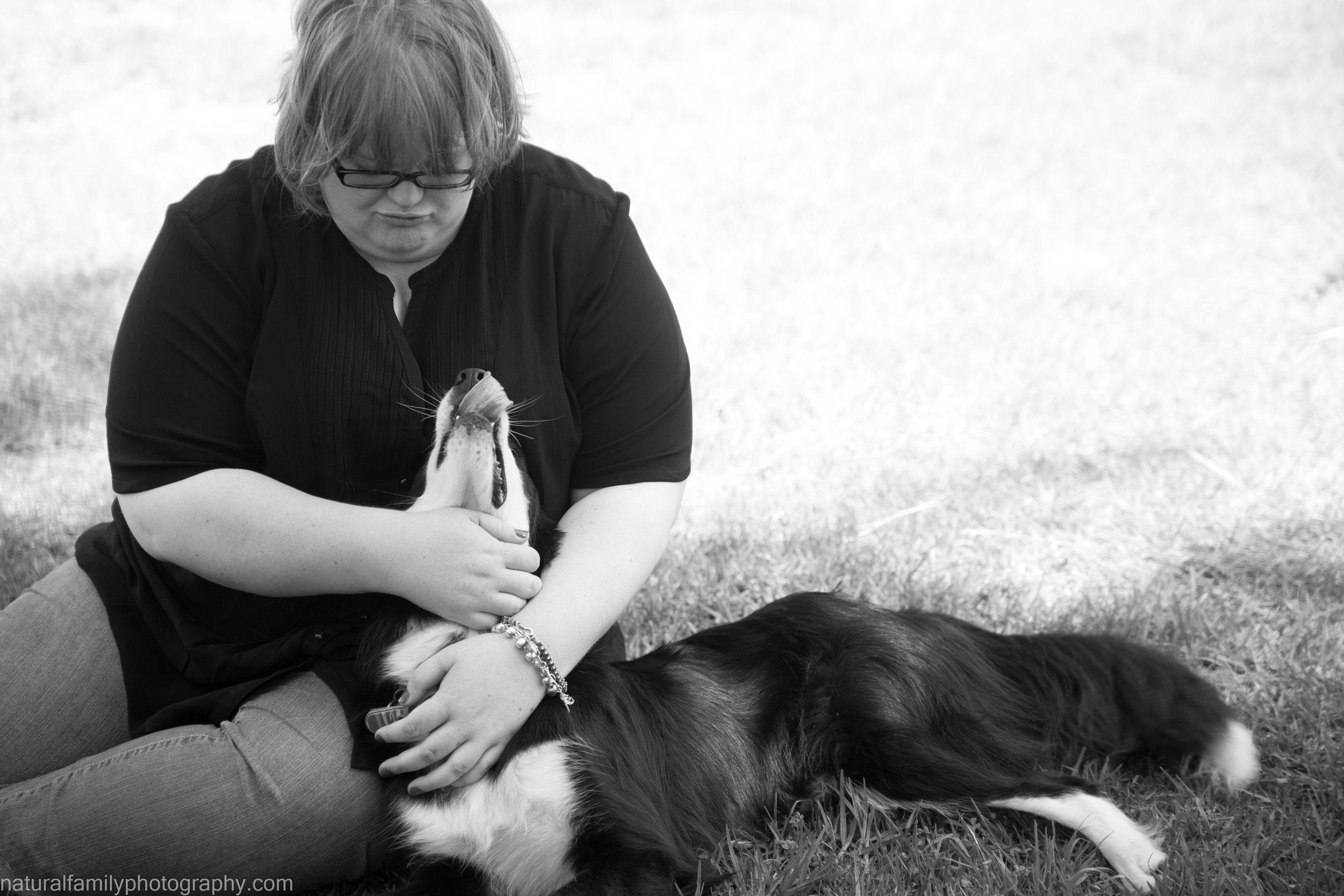 A person with glasses and curly hair sitting on grass, holding a dog’s head up, with the dog lying down and yawning.