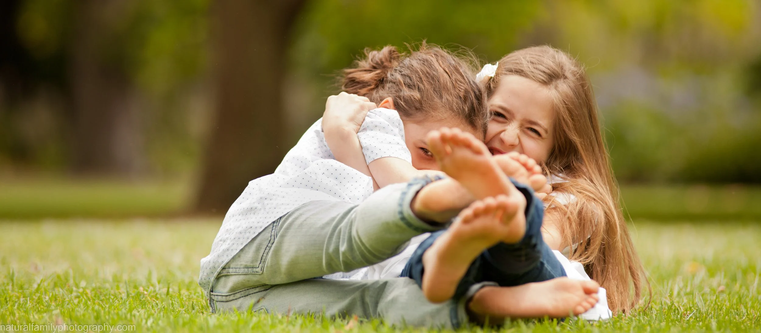 Two young girls playing on grass, one girl pinning the other down in a playful embrace, outdoor setting with trees in the background.