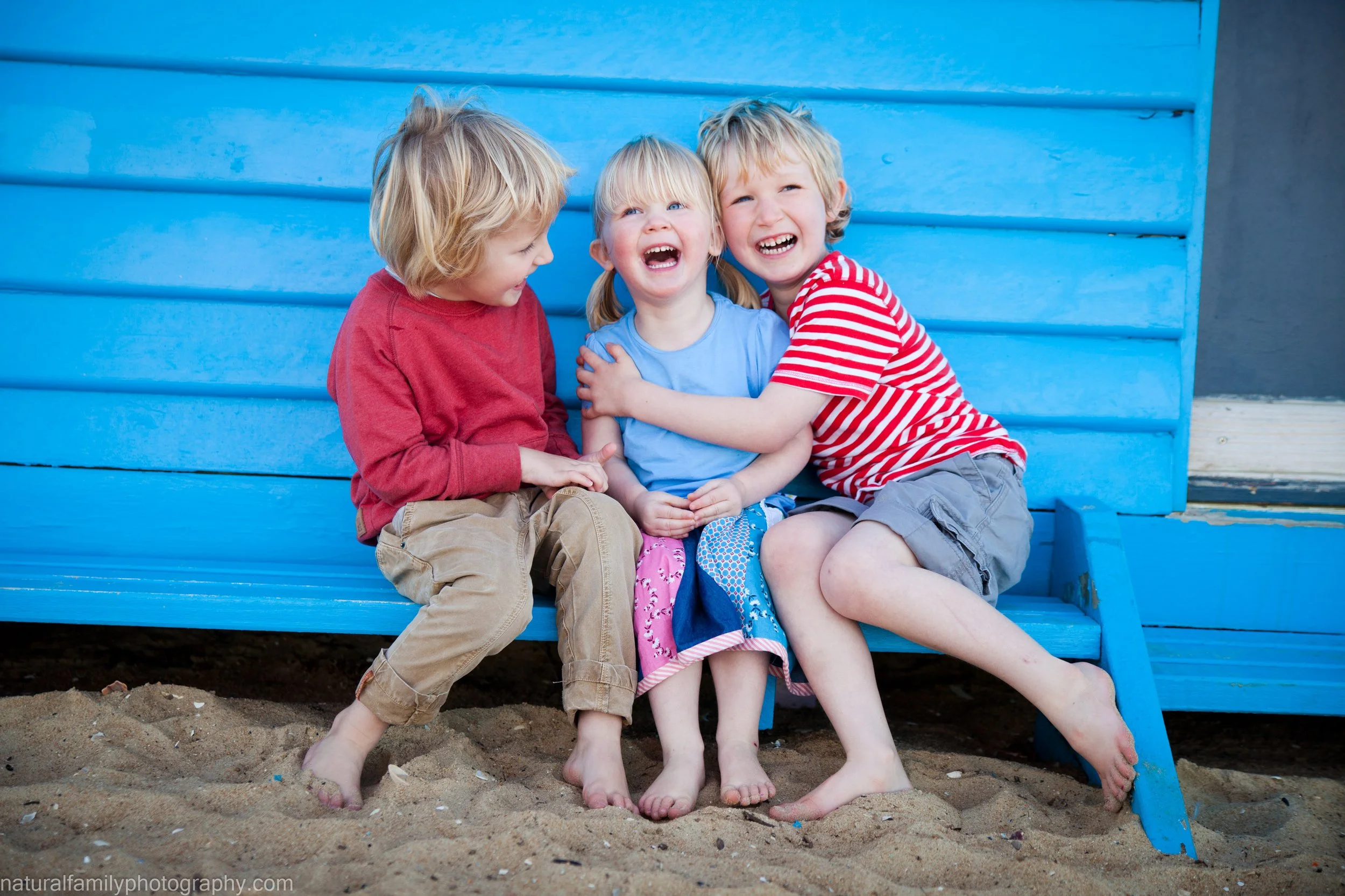 Three young children sitting on a blue park bench, laughing and hugging each other.