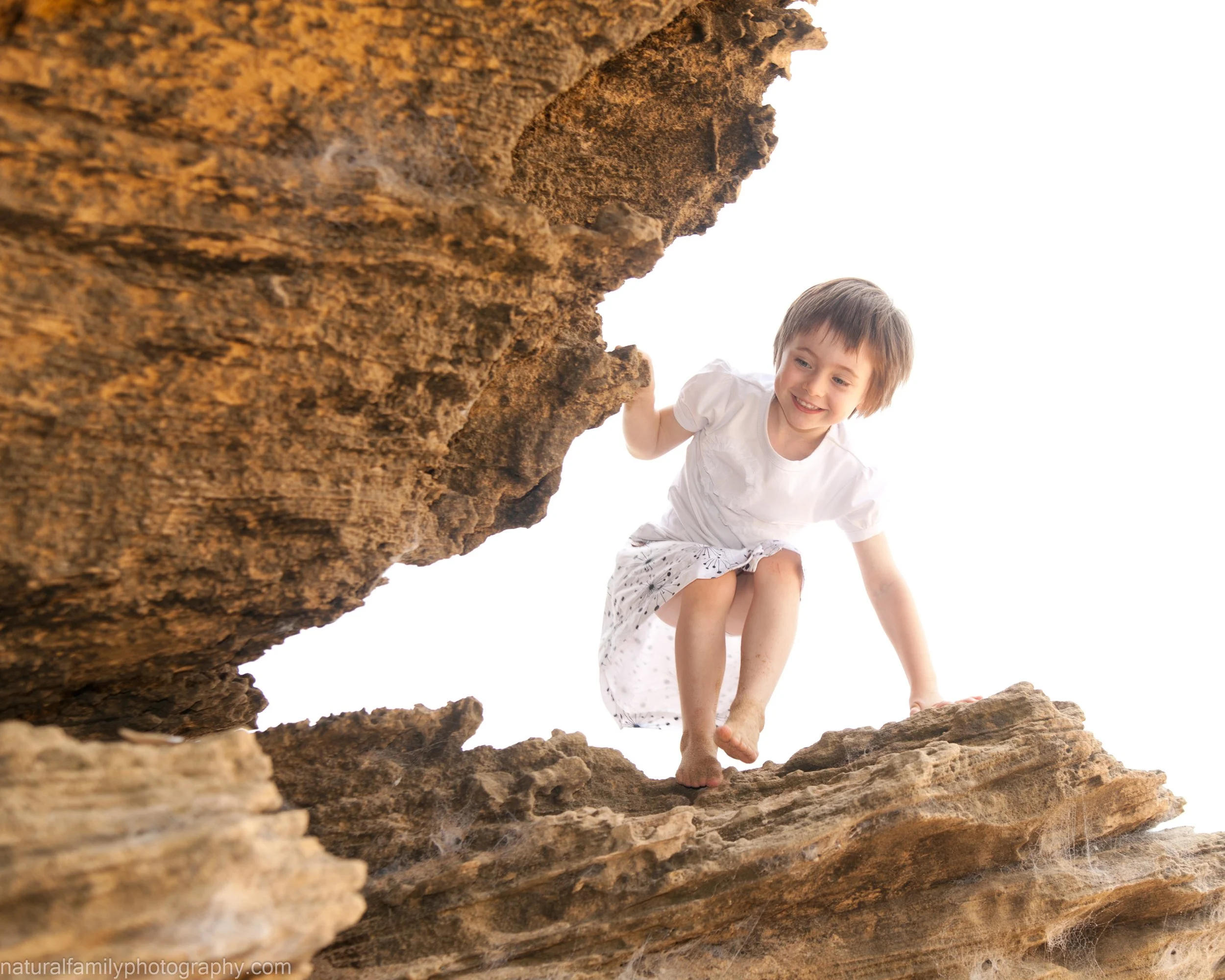 A young girl with short brown hair, wearing a white dress, is climbing on large brown rocks outdoors against a bright sky. Location portrait by Natural Family Photography, Melbourne.