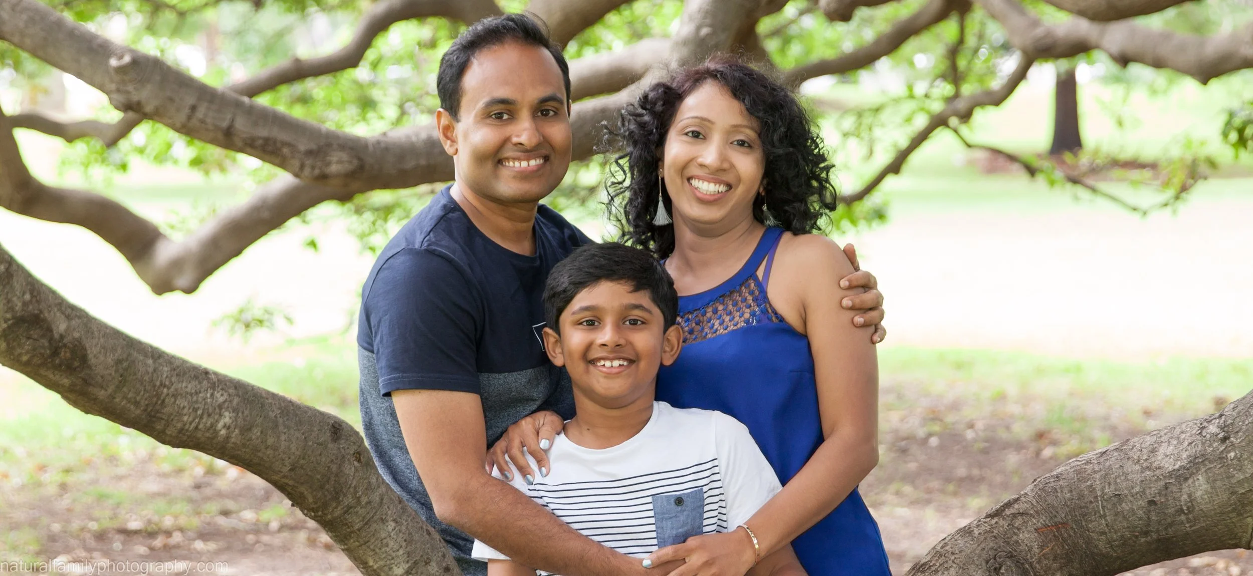 A family of three, a man, a woman, and a young boy, smiling and standing close together outdoors in a park with green leaves and tree branches in the background.