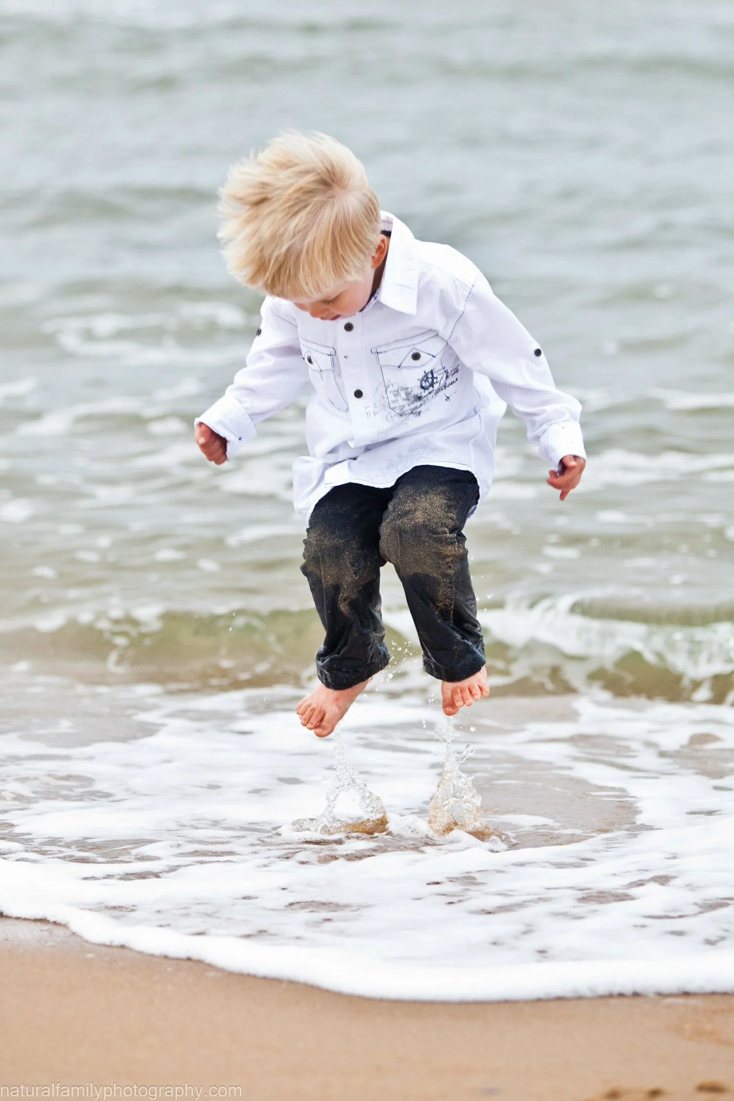 A young boy with blonde hair wearing a white shirt and black pants jumping in the shallow water at the beach.