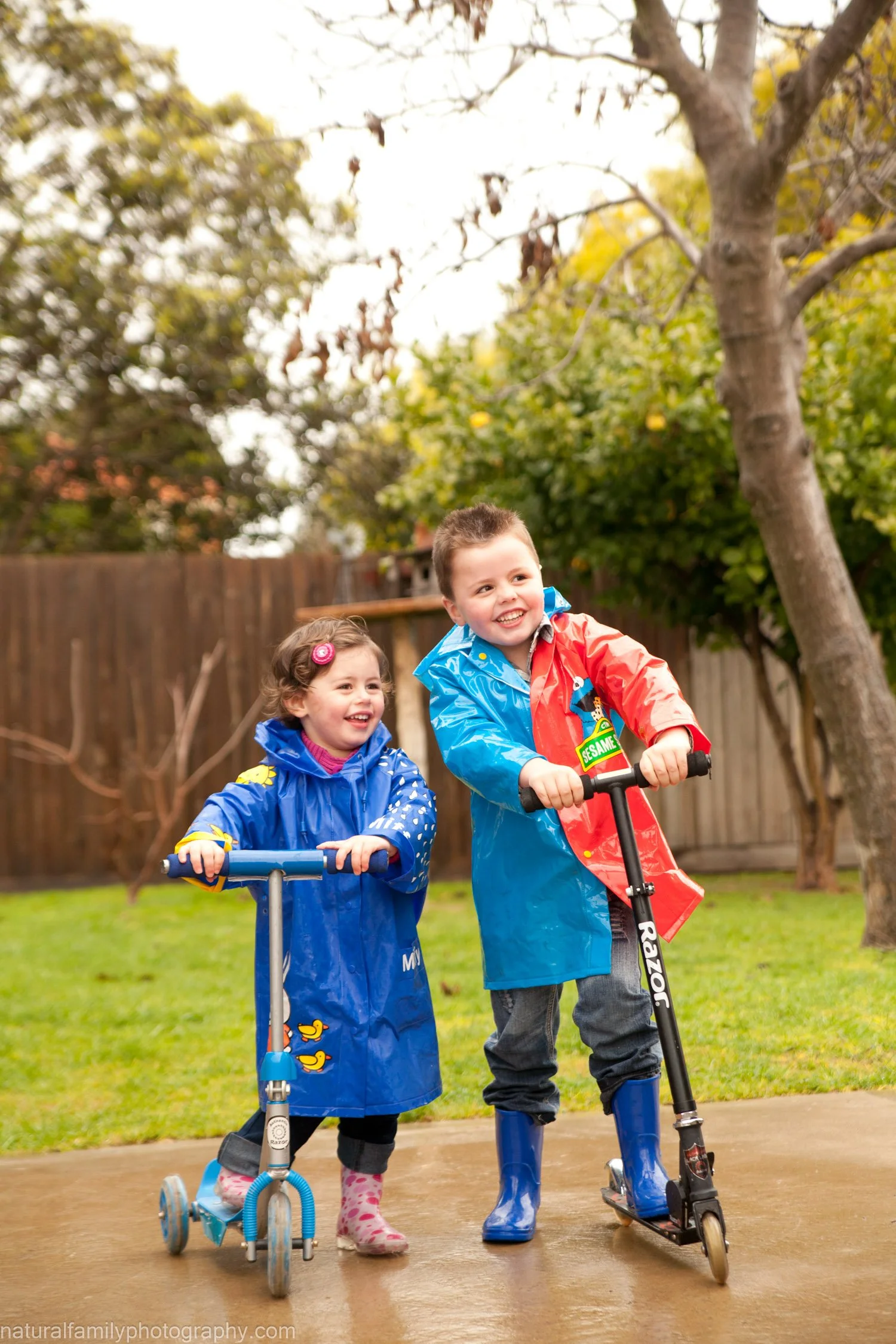 Two young children, a boy and a girl, playing outside on scooters. They are smiling and wearing colorful raincoats and rain boots, with trees and a wooden fence in the background.