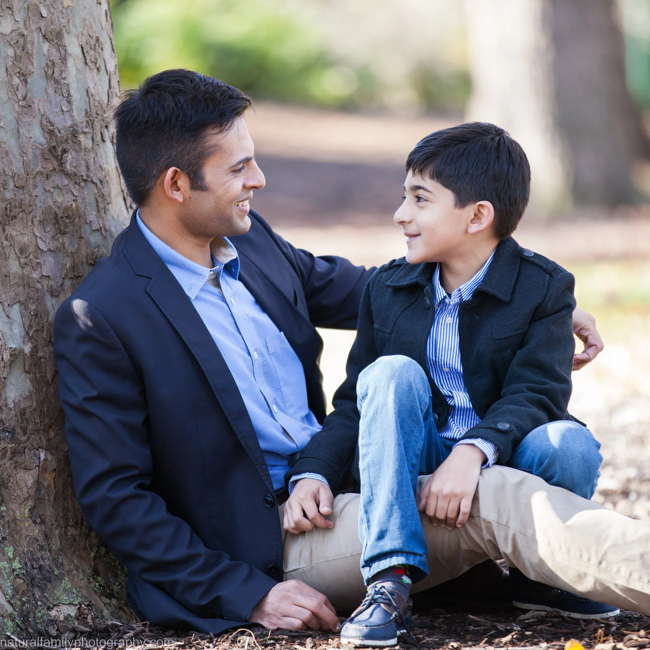 A man and a boy sitting on the ground outdoors, smiling at each other, with trees in the background.