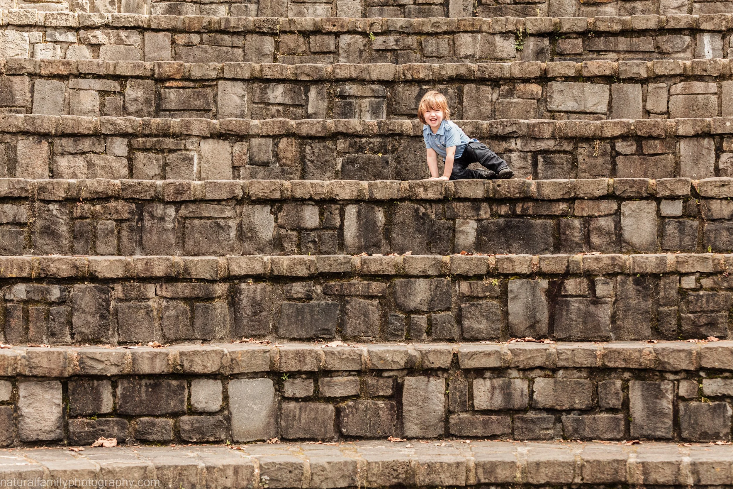 A young boy with blond hair, wearing a blue shirt and black pants, climbing on a stone staircase and smiling.