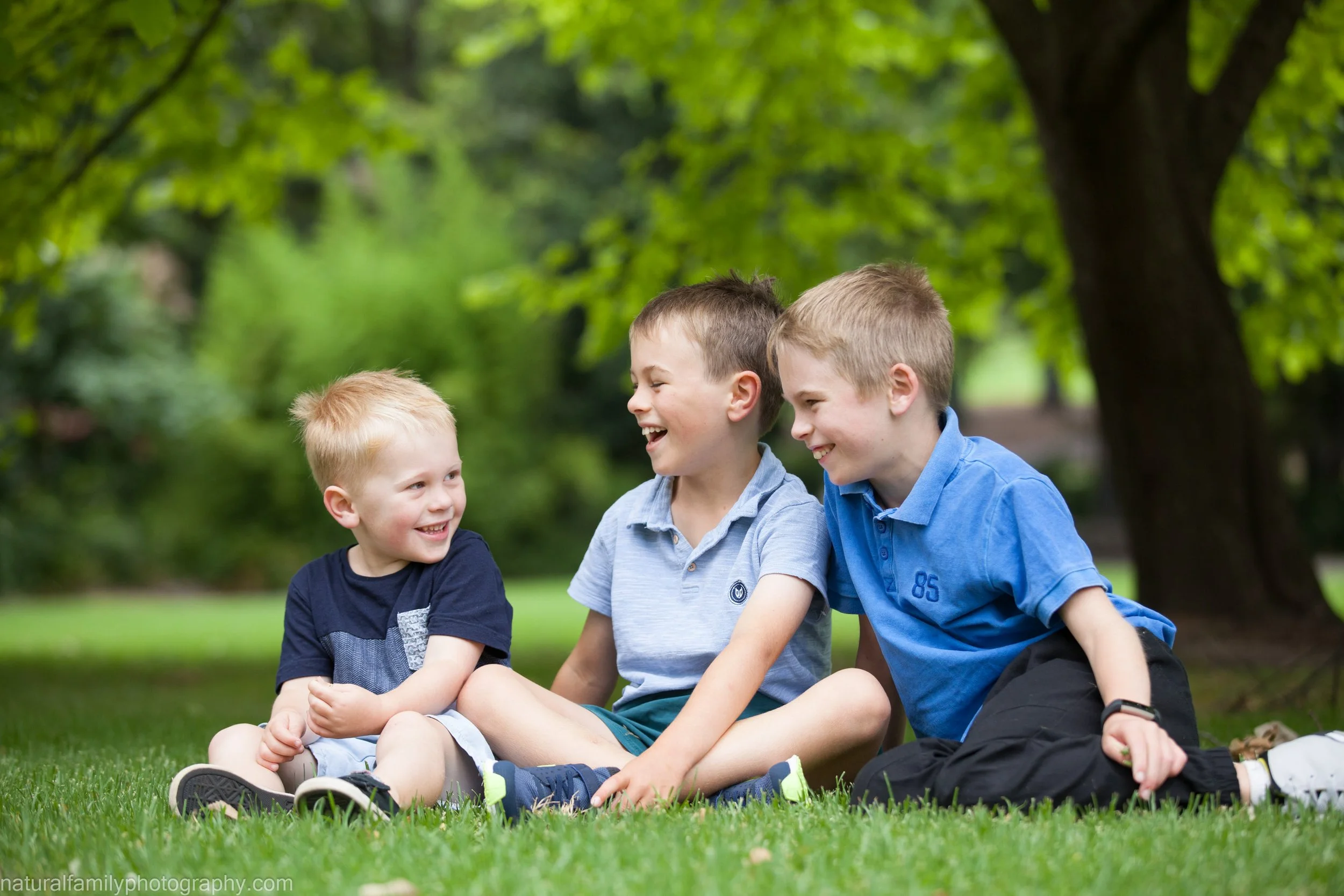 Three boys sitting on grass in a park, laughing and playing together.