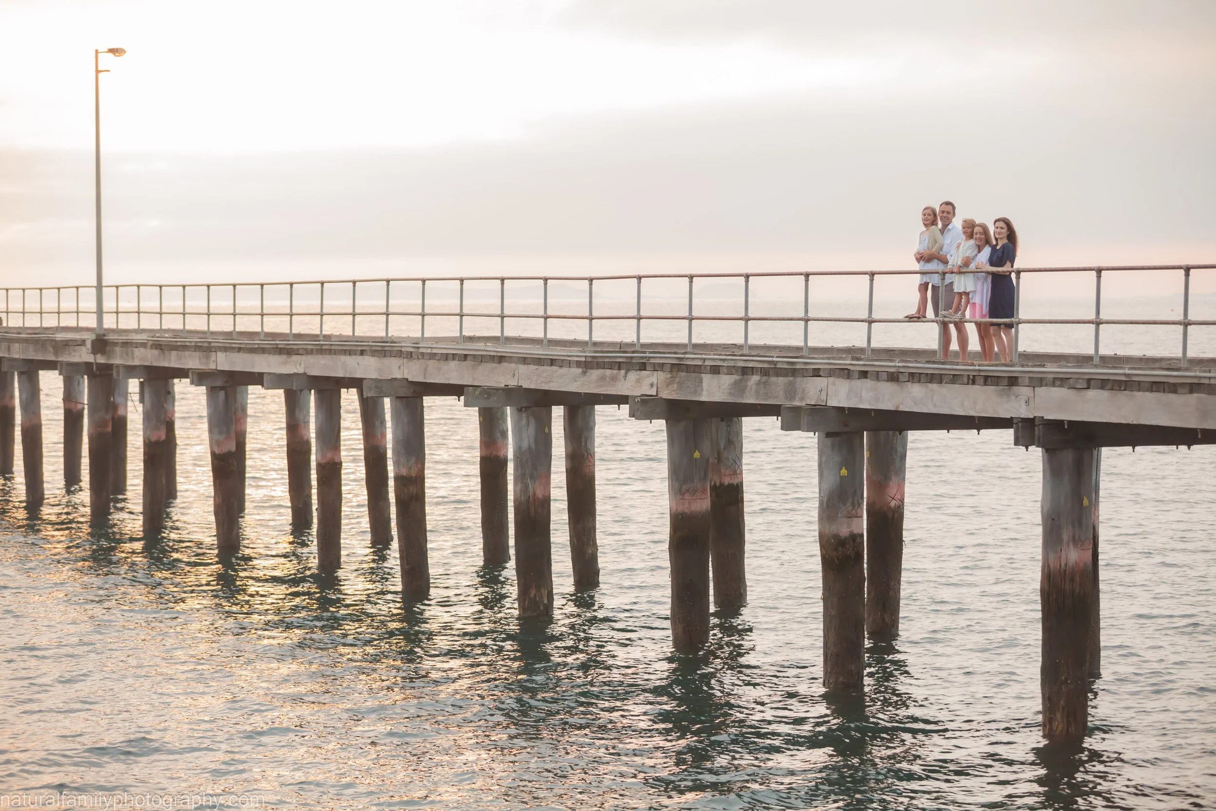 A group of five people standing on a pier by the water at sunset, looking out over the ocean.