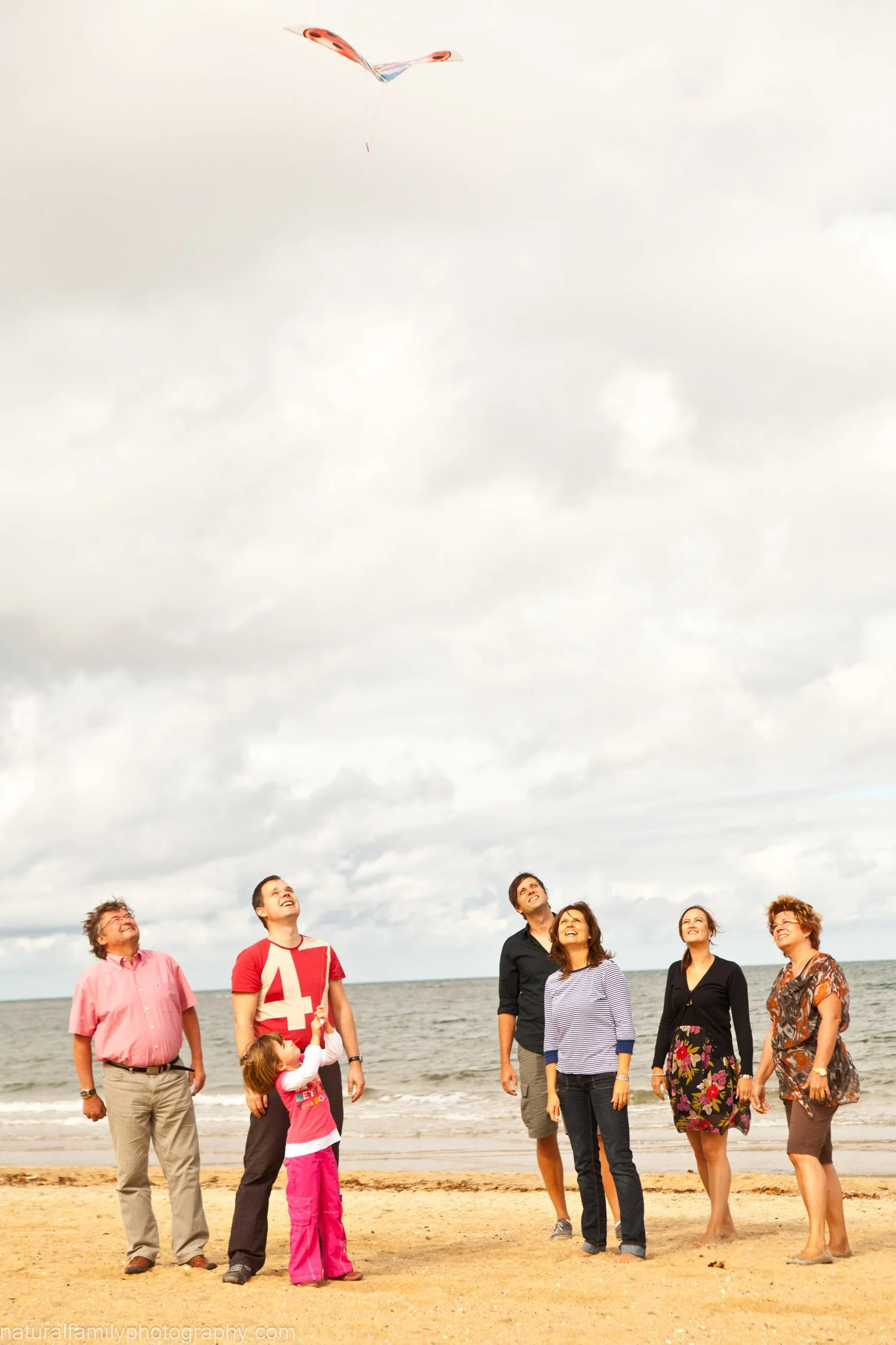 A group of seven people, including children and adults, standing on a beach, flying a kite in the cloudy sky.