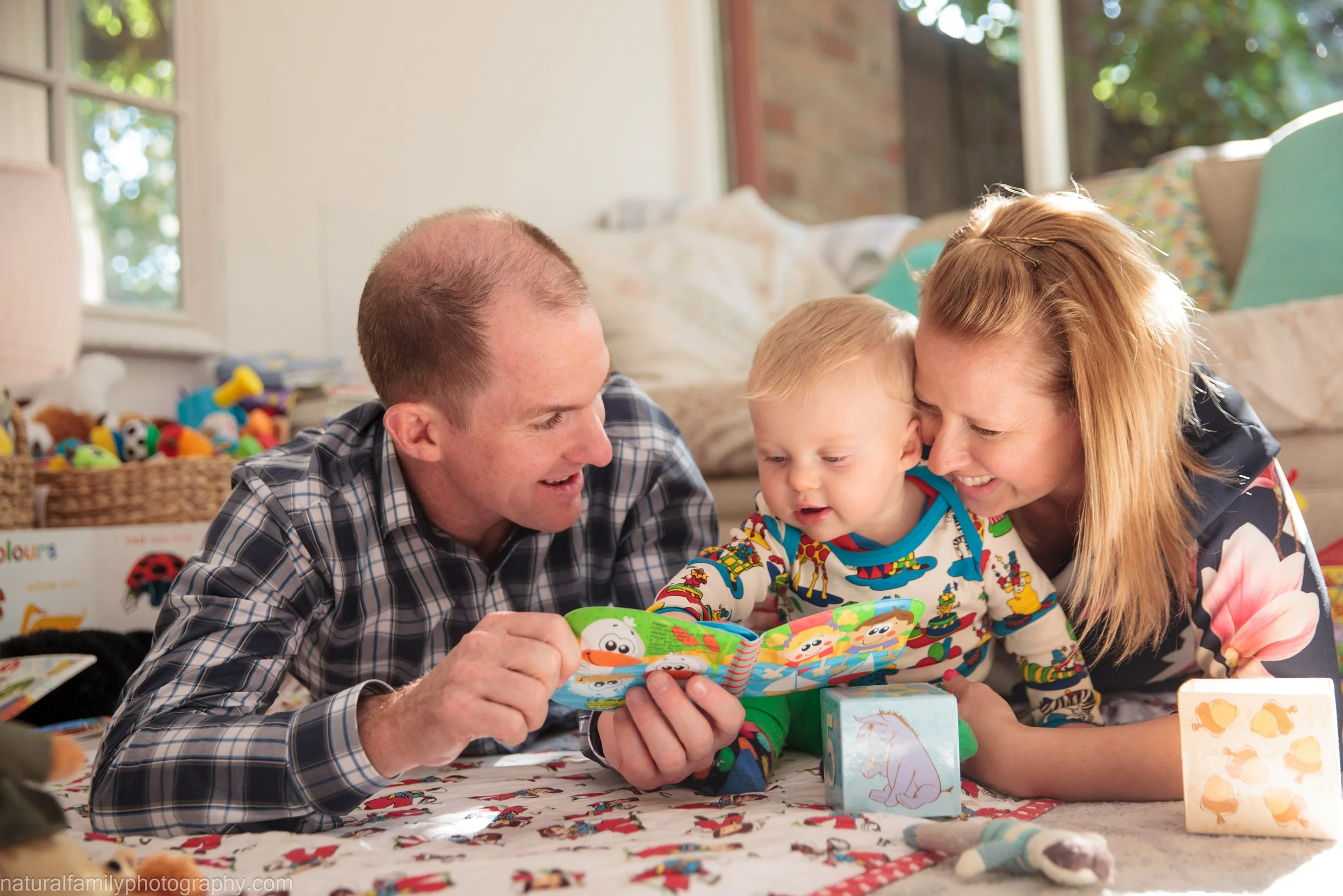 A family of three, a father, mother, and young child, lying on the floor and looking at a colorful book together in a cozy, sunlit room filled with toys and stuffed animals.