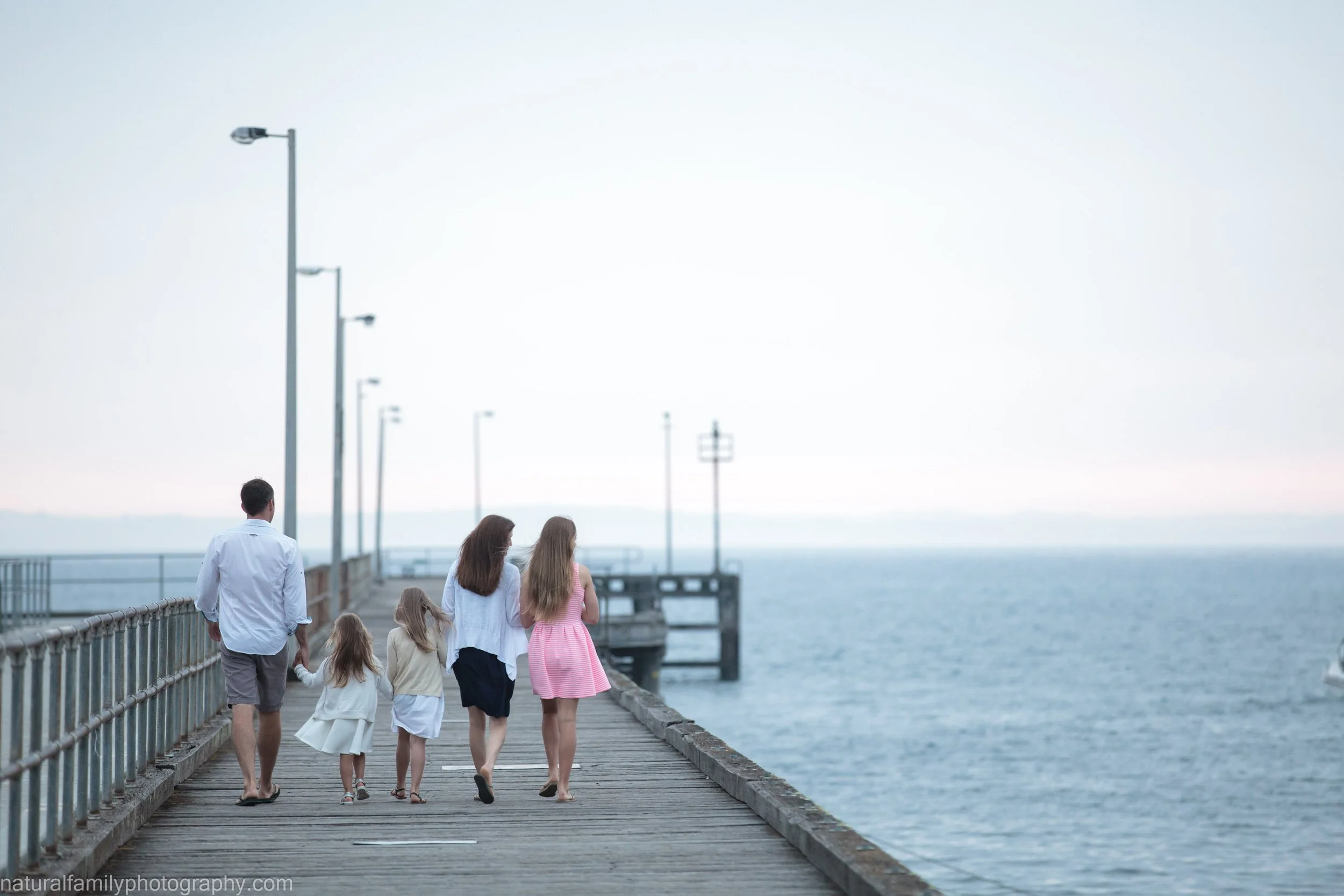 A family of five walking on a wooden pier by the sea with lampposts along the side, some adults and children holding hands, and the sky and water in the background.