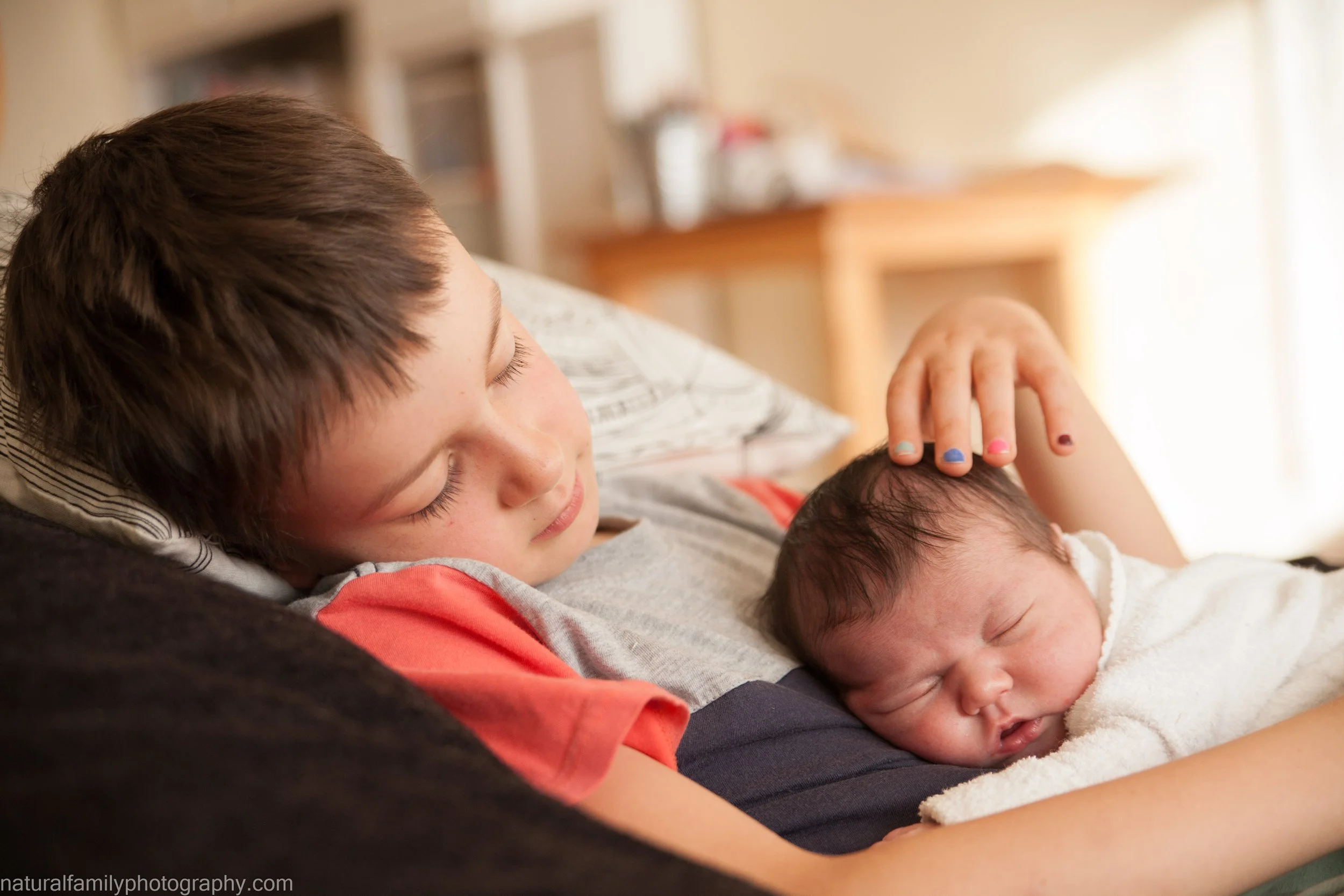 A young boy with short brown hair gently cradles a newborn baby, lying on his chest, while resting on a pillow in a warmly lit room.