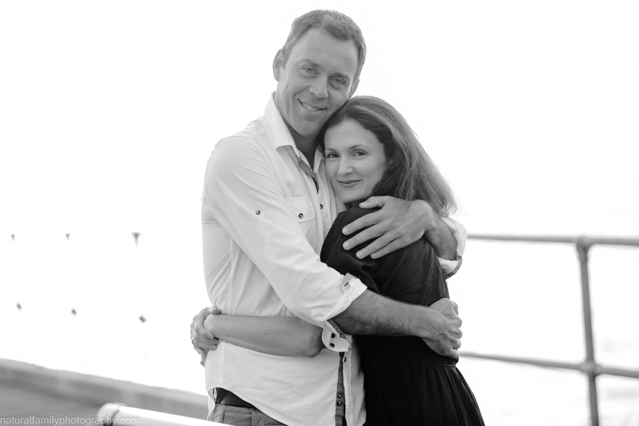 A man and woman hugging each other outdoors, smiling at the camera in black and white.