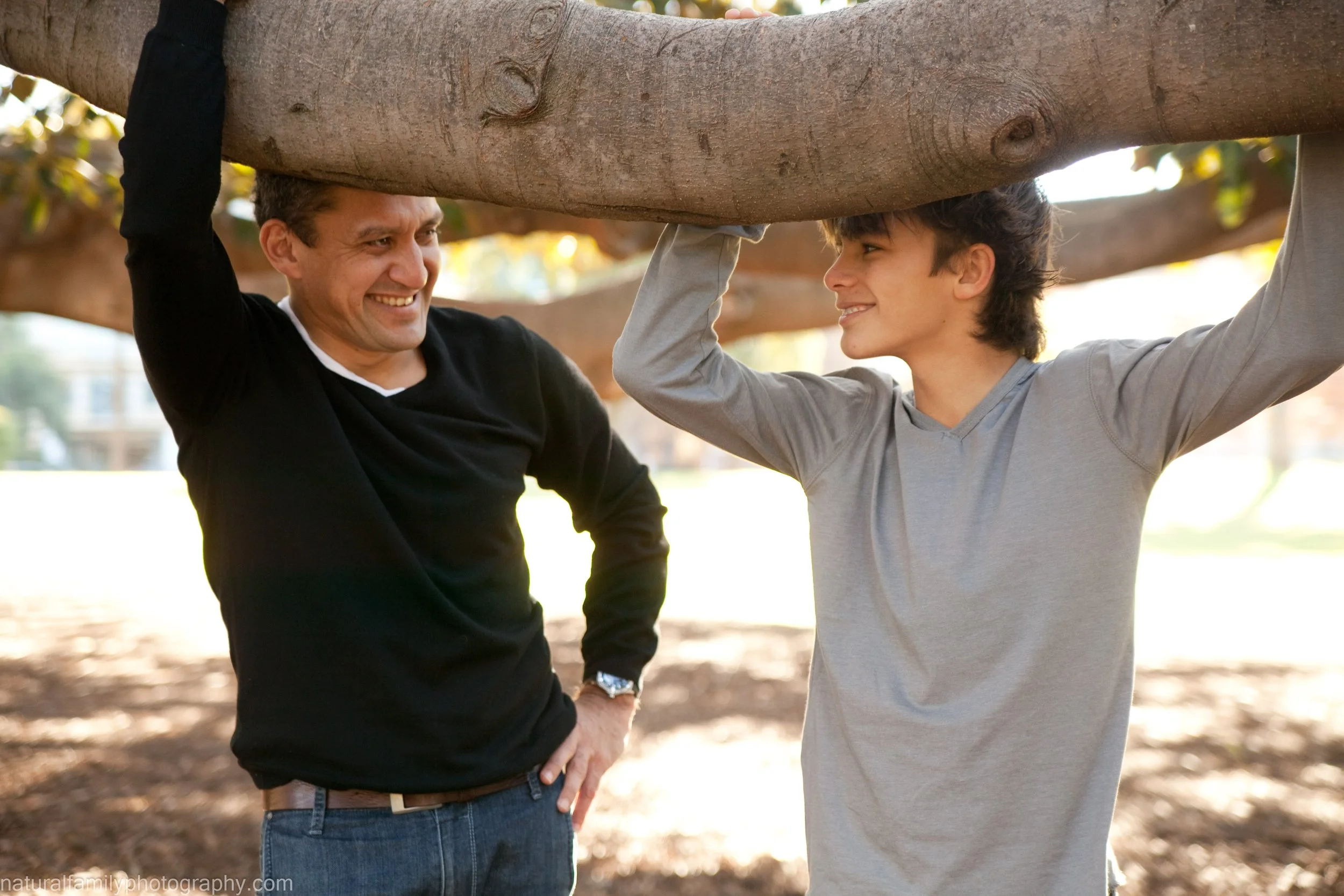 A man and a teenage boy smiling and talking while holding up a large tree branch in a park.