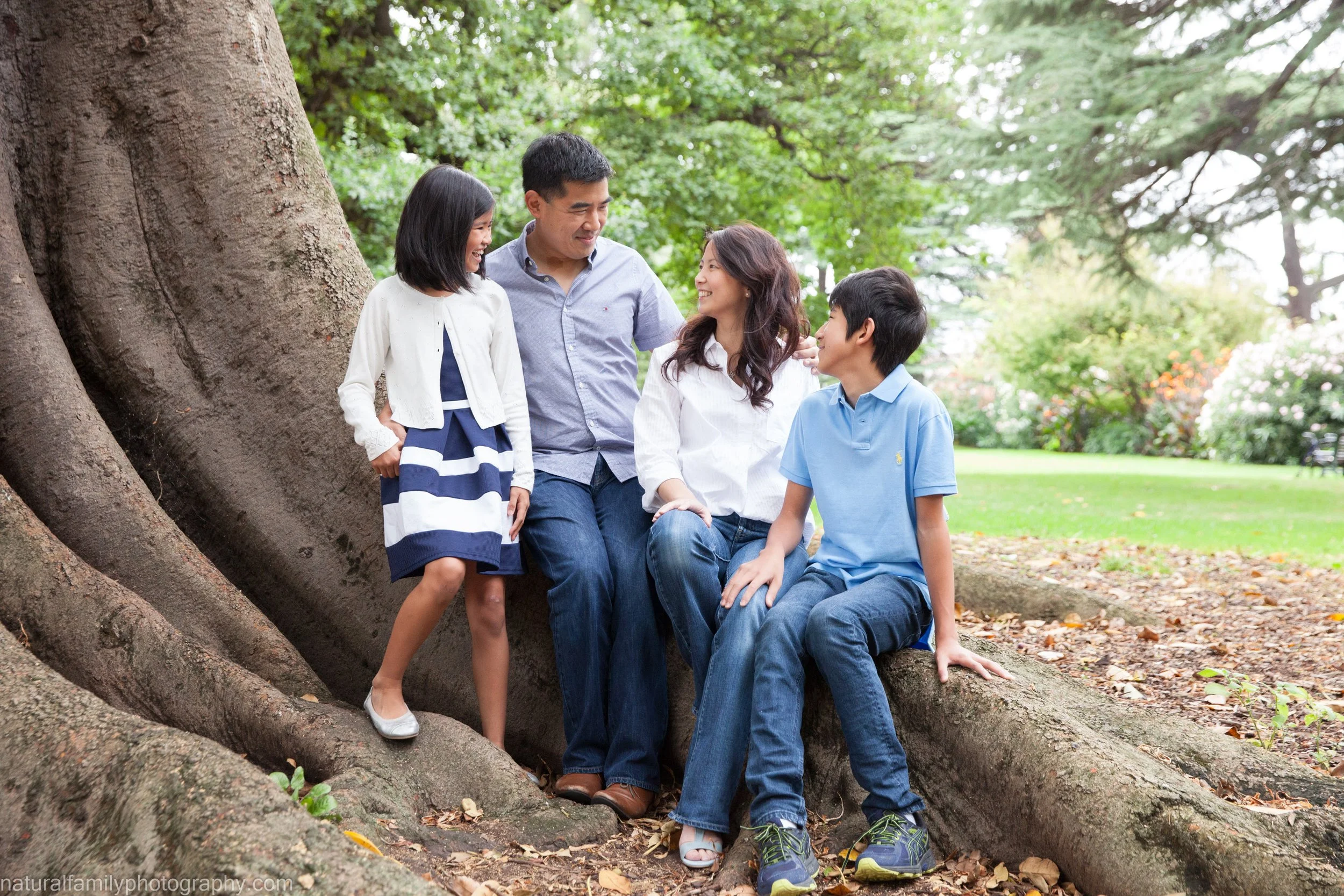 A family of five sitting and standing by a large tree in a park, smiling and engaging with each other.
