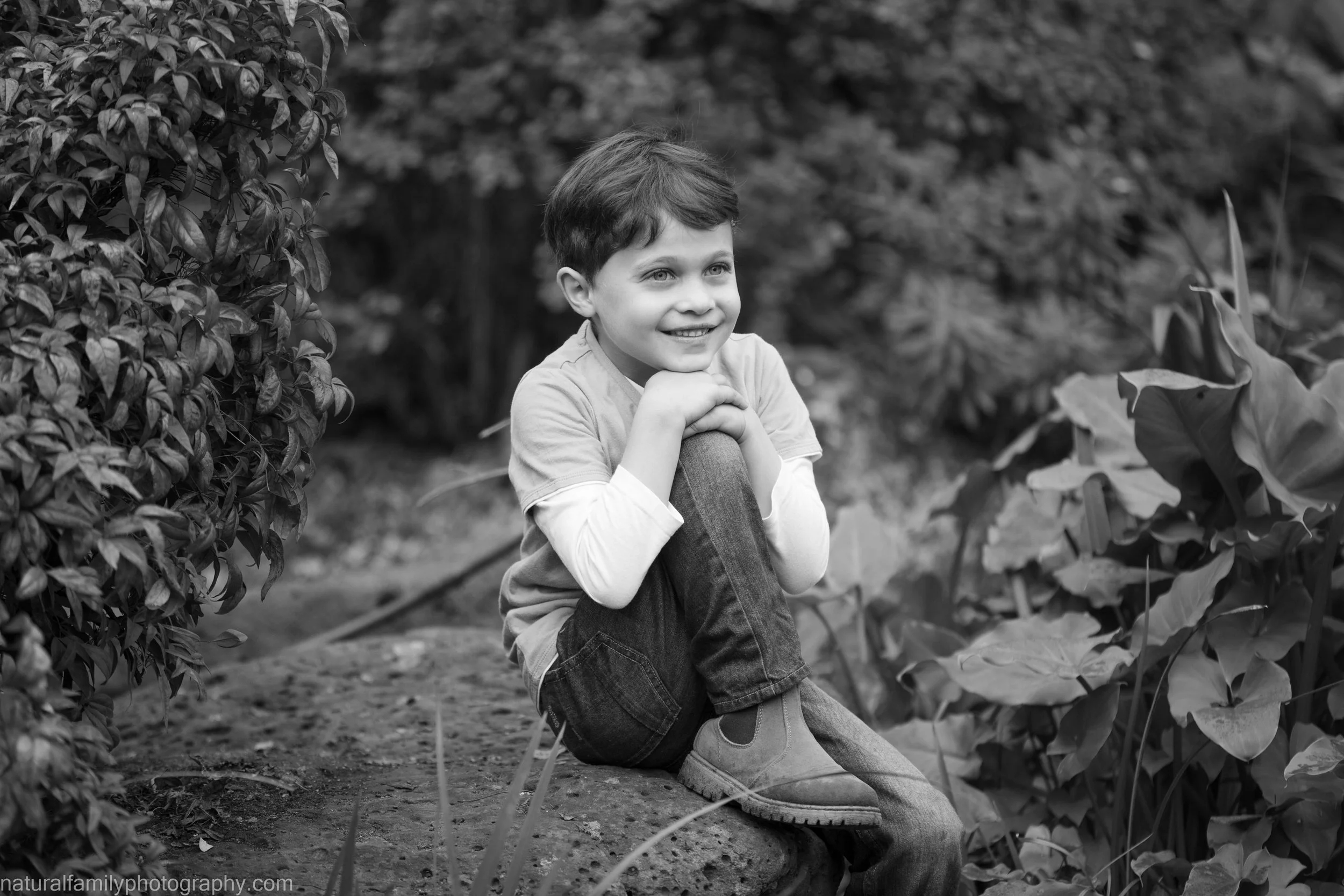 A young boy sitting on a stone in a garden, smiling and resting his chin on his hands, surrounded by plants and foliage.