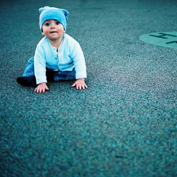 A baby in a light blue hat and jacket crawling on a textured greenish-blue playground surface. Photographed using traditional analogue film and a vintage lens by Natural Family Photography, Melbourne.