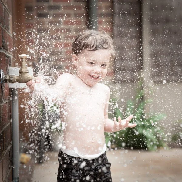 A young boy playing outside and splashing water from a faucet, smiling and shirtless. Joyful portrait by Natural Family Photography, Melbourne.