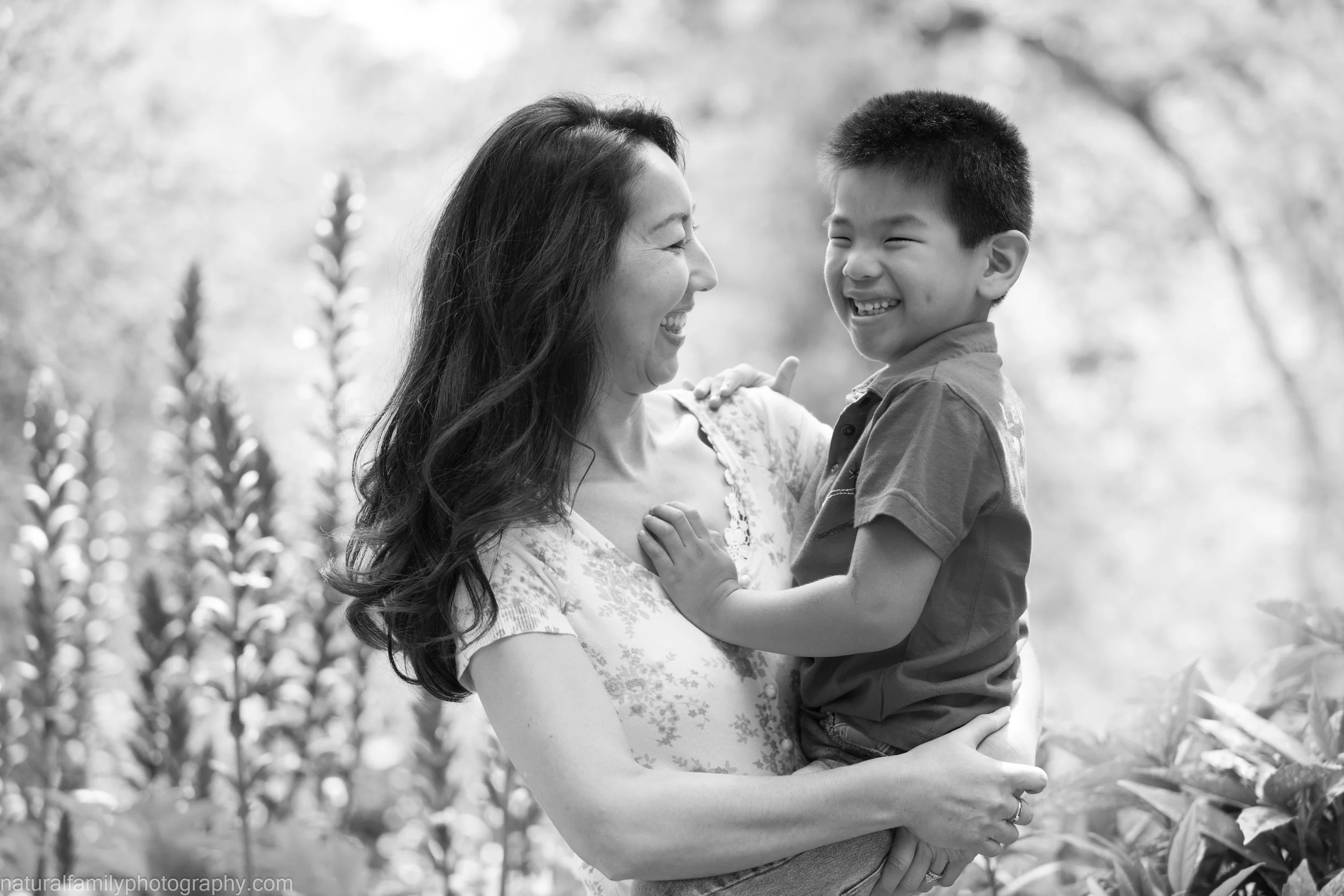 A woman holding a young boy in an outdoor setting, both smiling and facing each other.