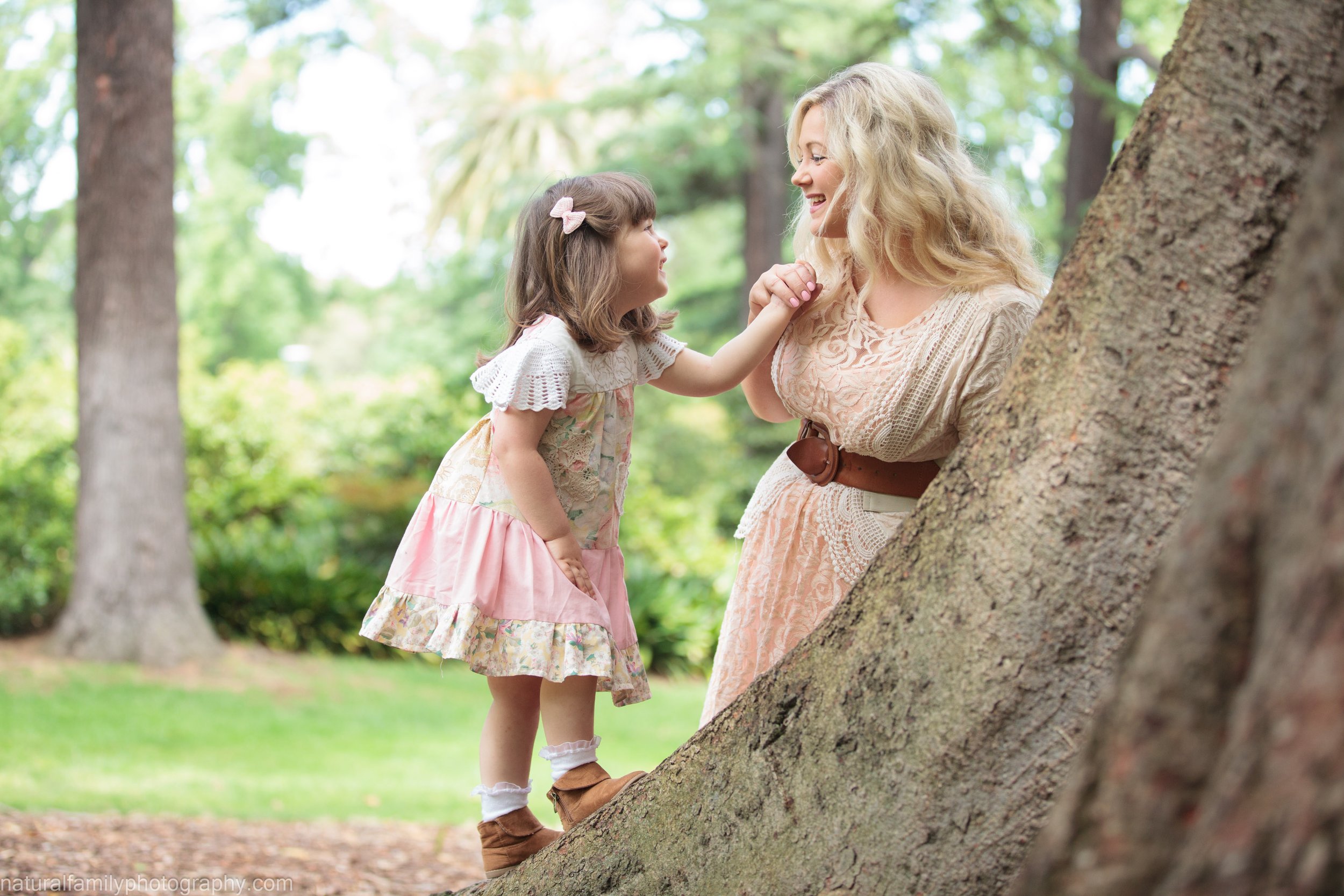 A young girl and woman smiling and holding hands, standing behind a tree in a park with green trees and grass.
