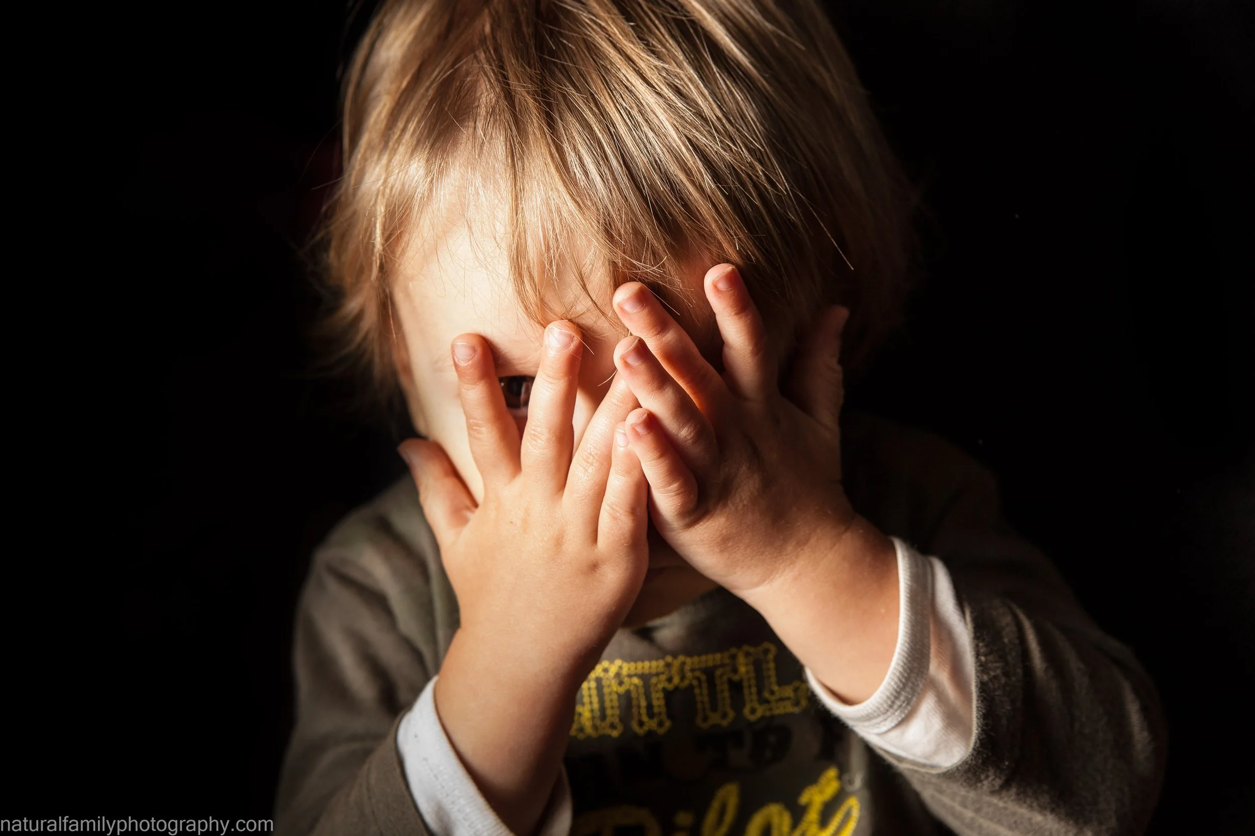 A young child with light brown hair covering part of their face, peering through their fingers against a dark background. Creative studio portrait by Natural Family Photography, Melbourne.