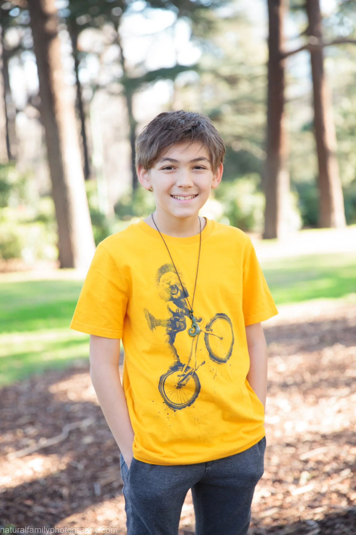 A smiling boy wearing a yellow T-shirt with a bicycle graphic and a black necklace, standing outdoors in a park with trees and green grass in the background.