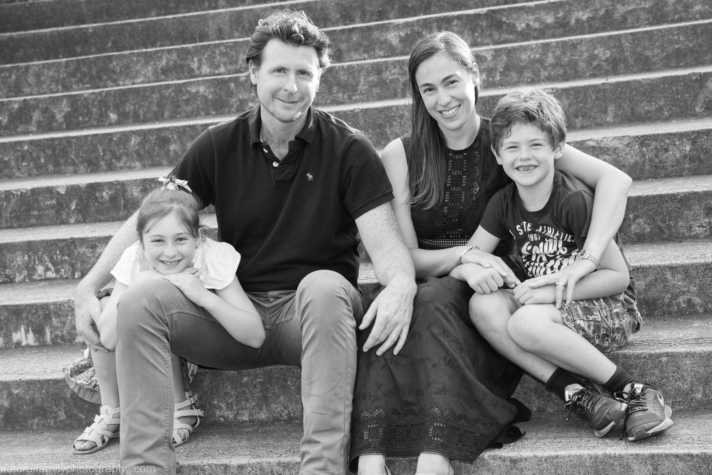 A family of four sitting on concrete stairs outdoors, smiling for the camera in black and white.