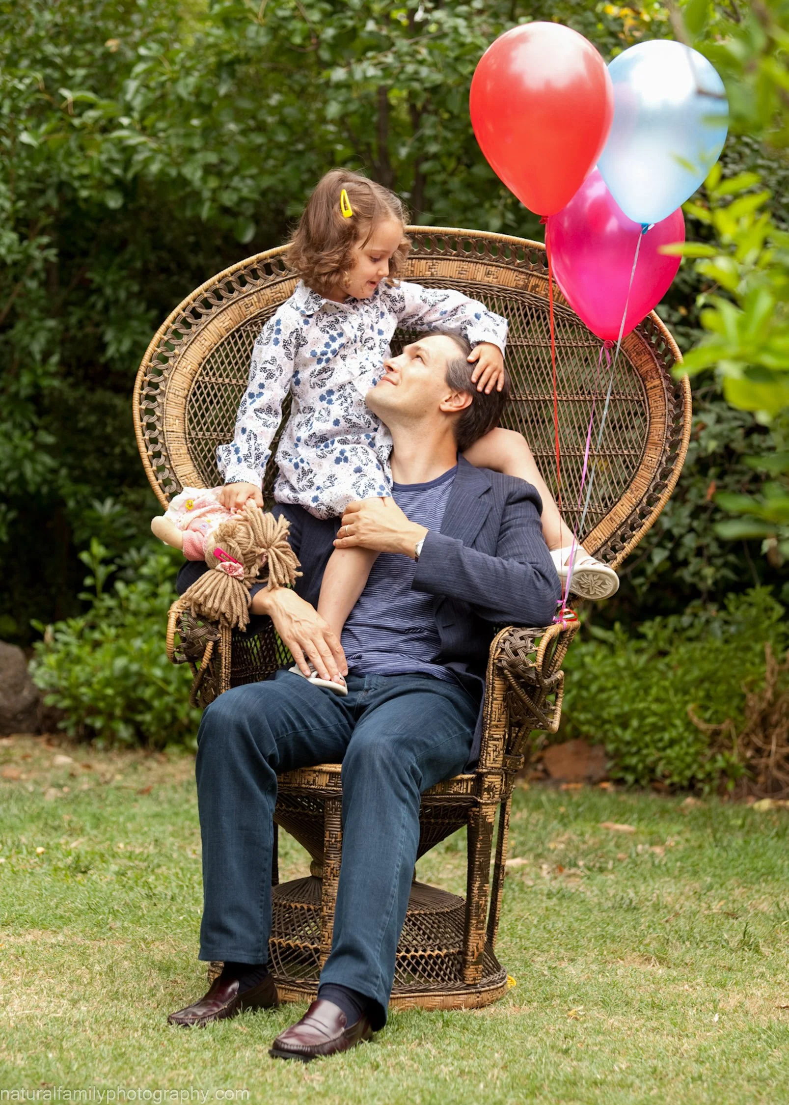 A man and a young girl are sitting on a wicker chair outdoors, with the girl sitting on the man's lap. The girl is leaning towards the man, who is looking up at her. Three colorful balloons are attached to the chair. The background is filled with gre