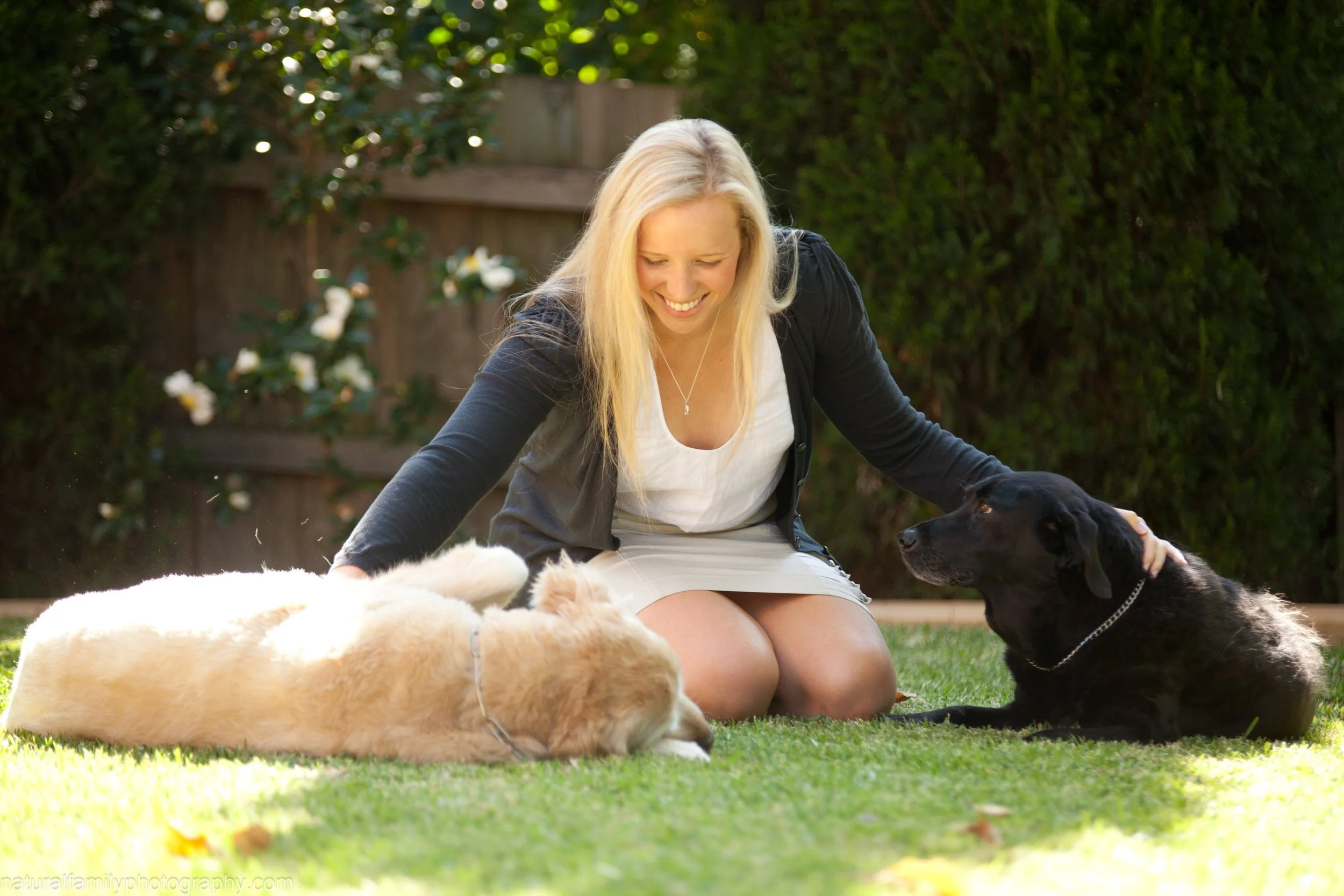 A woman with long blonde hair sitting on the grass outside, smiling, petting a black dog and a golden retriever puppy lying on the grass.