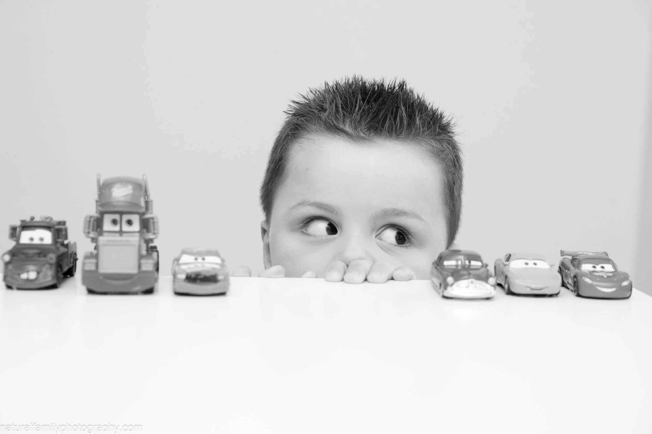 A young boy with spiked hair peeking over a table with toy cars lined up in front of him, looking to the side.