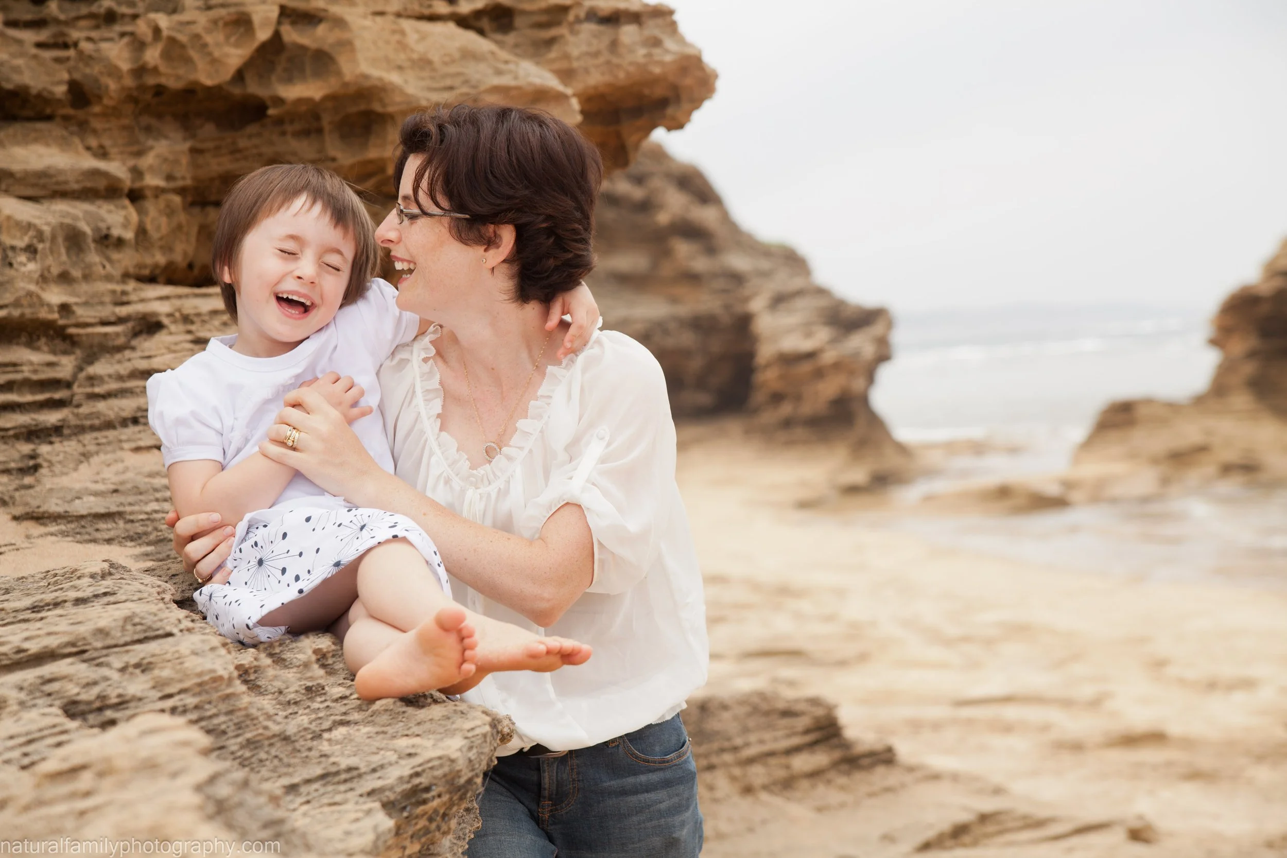 A woman and a young girl sharing a joyful moment on a rocky beach, with the woman cuddling the girl and both smiling. Location coastal portrait by Natural Family Photography, Melbourne.
