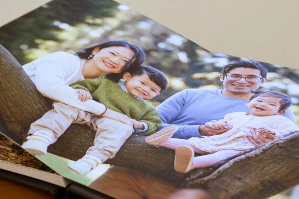 Archival, hand-bound album featuring photography of a family of four sitting on a tree branch outdoors, smiling at the camera. The family includes a woman, a young boy, a man, and a baby girl. Made by Natural Family Photography.