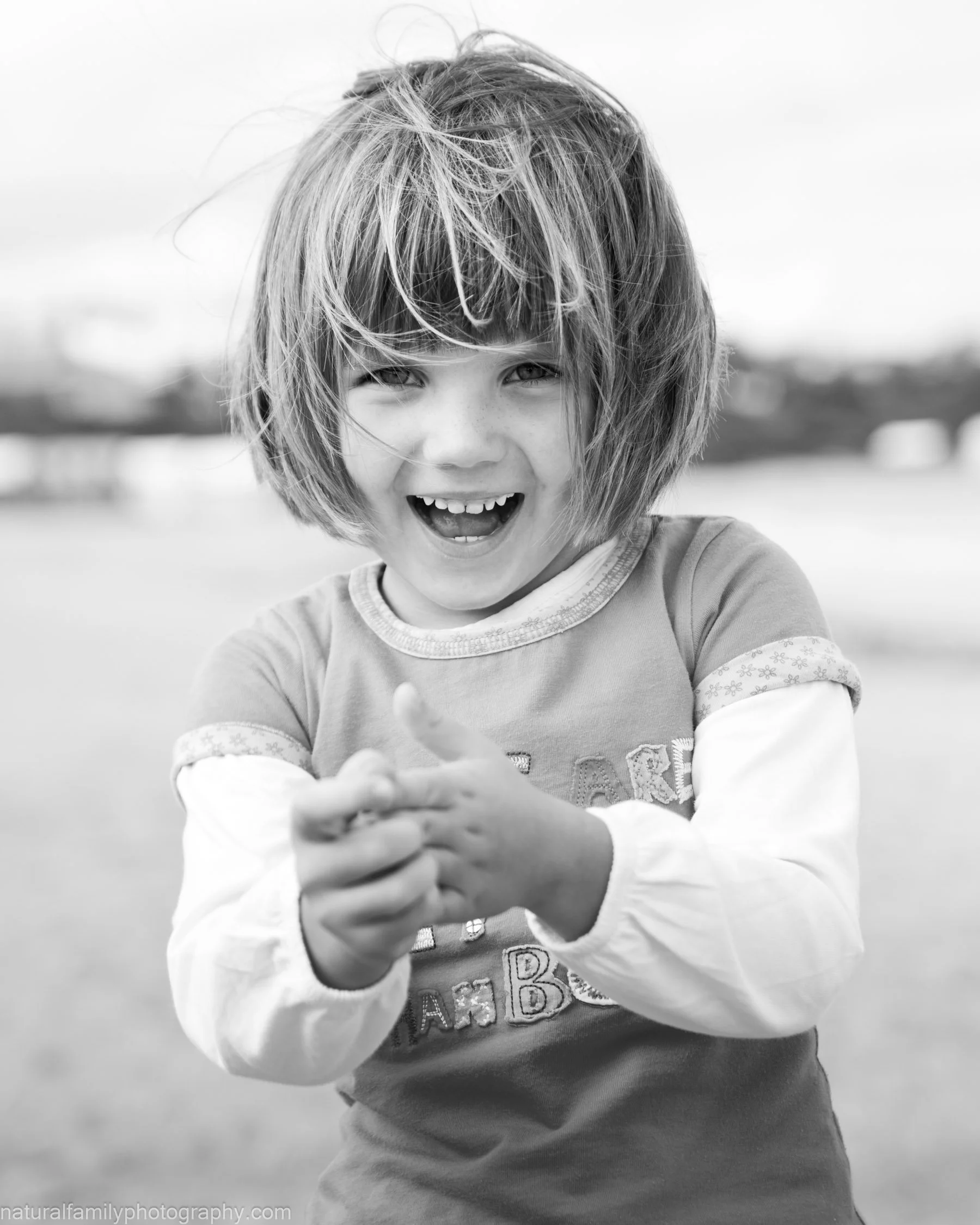 A young girl with messy hair smiling and clapping outside in black and white.