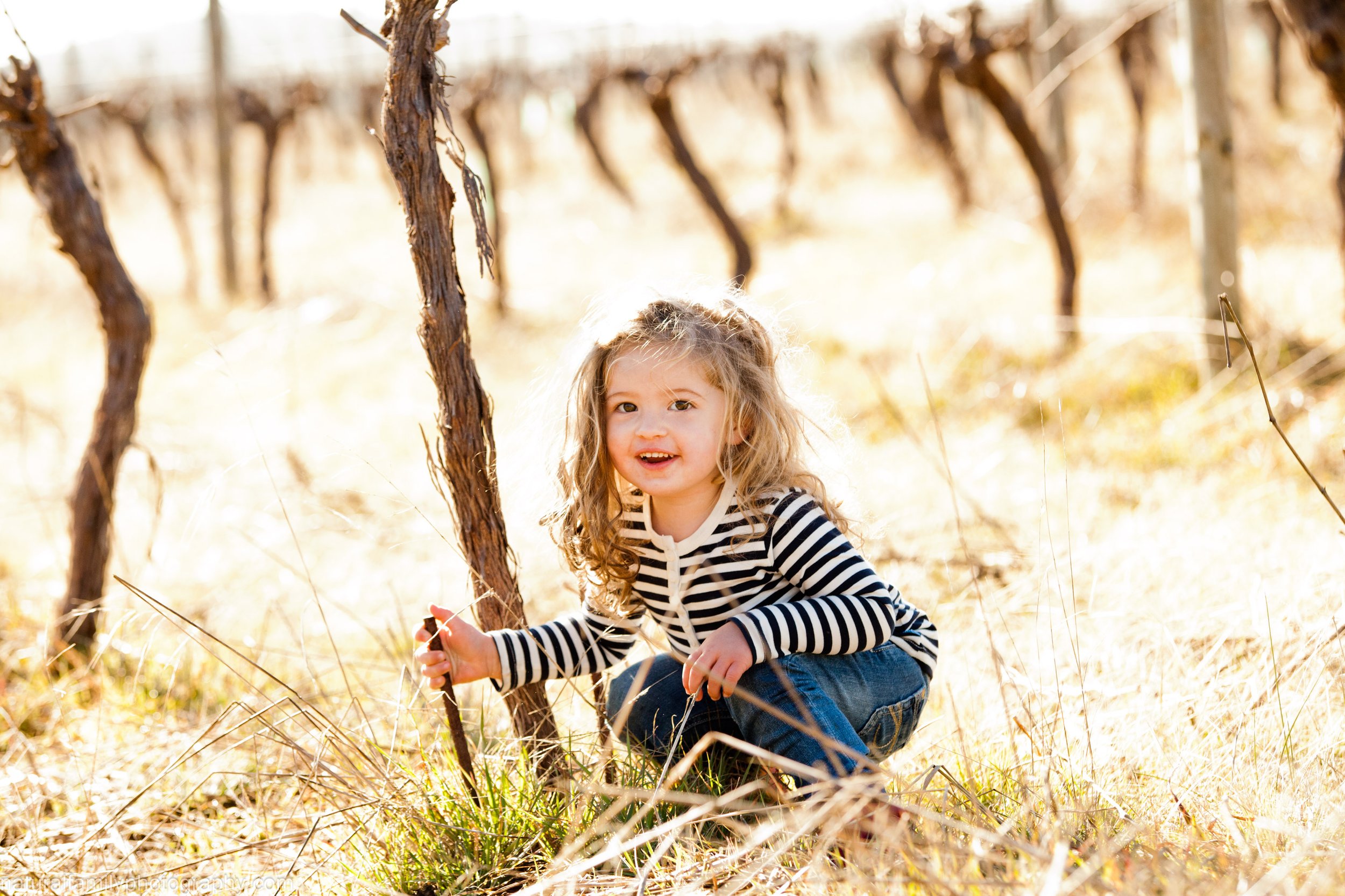 A young girl with curly blonde hair, wearing a black and white striped shirt and jeans, crouches in a bright, sunlit field among dry grass and twisted vines, holding a small stick and smiling at the camera.